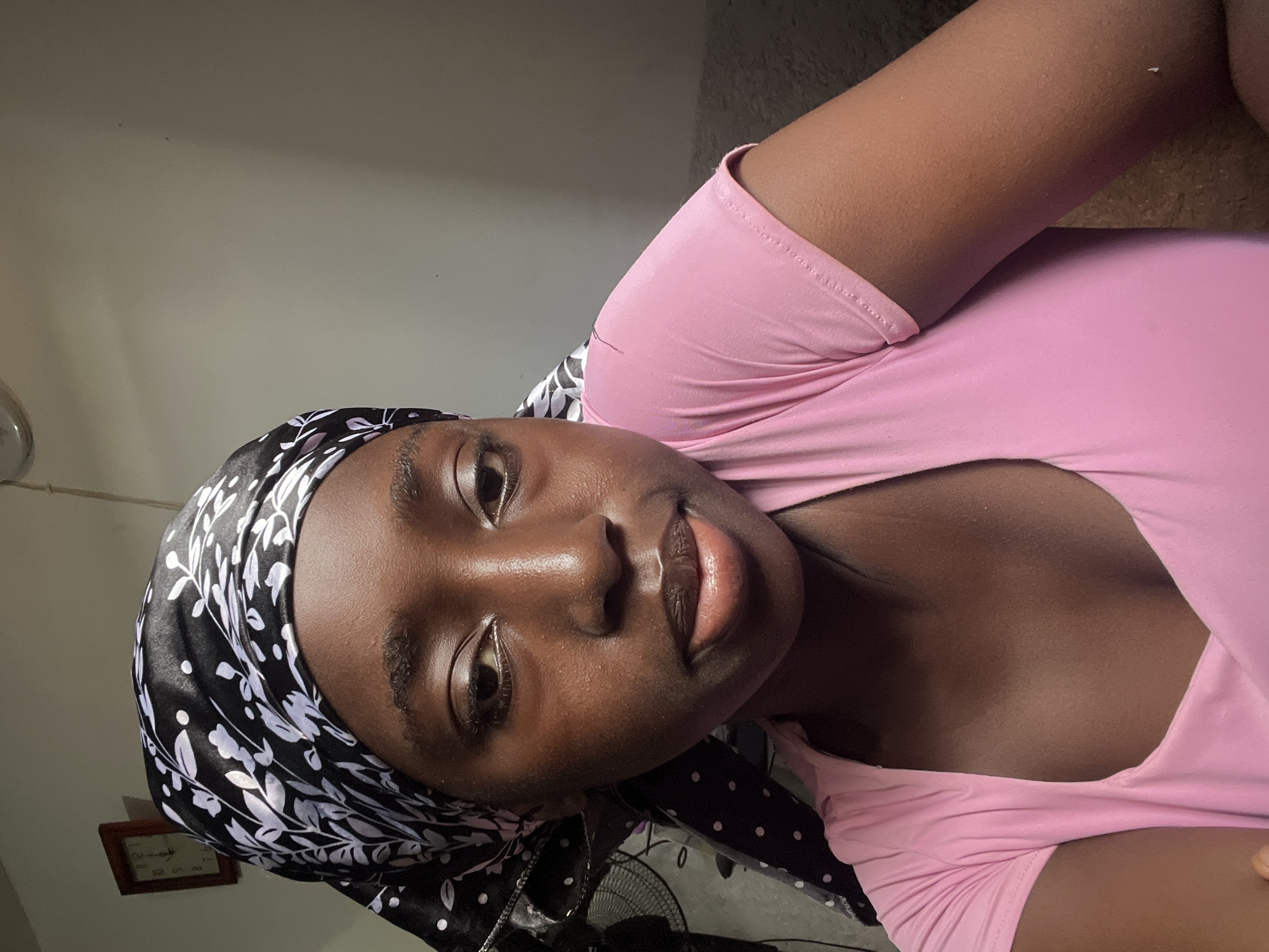 A young, dark-skinned woman wearing a pink shirt and a black and white headscarf is depicted in an eye-level, close-up shot. She appears to be indoors against a plain, light-colored wall. In the background, there is a framed picture, a ceiling fan, and a standing fan. The location is unknown.