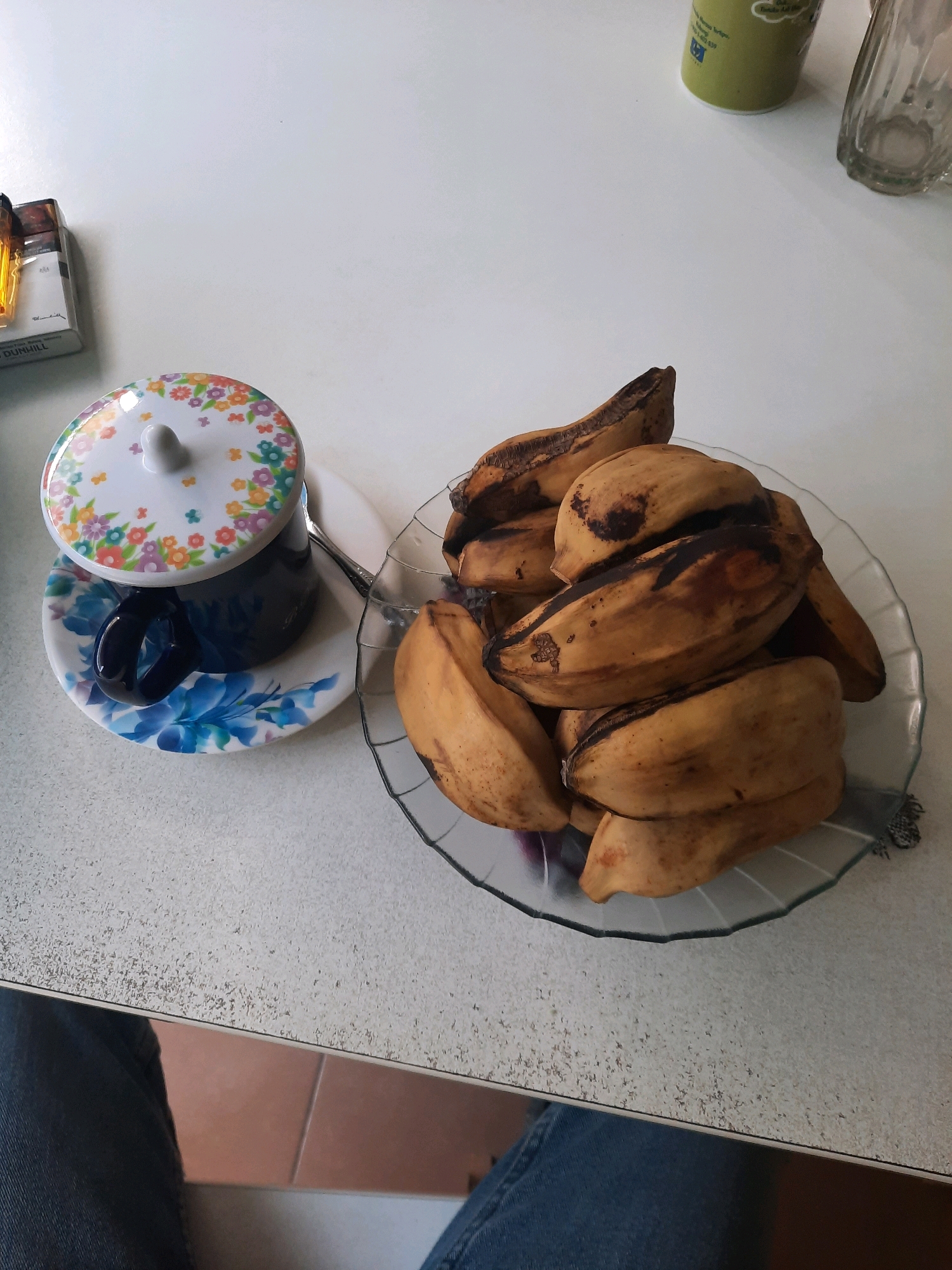 This image depicts a close-up, high-angle shot of a table in Babakan Madang, Indonesia. The table surface is light-colored with a speckled texture. On the left side of the table, a pack of Dunhill cigarettes is visible. To the right of the cigarettes, there is a dark blue coffee cup with a decorative floral lid, resting on a saucer with a blue floral pattern. A spoon is partially visible next to the cup. In the center-right of the image, a clear glass bowl contains several overripe bananas with brown spots. In the background, out of focus, a green container and a glass are visible. The bottom of the image shows a person's legs wearing blue jeans.