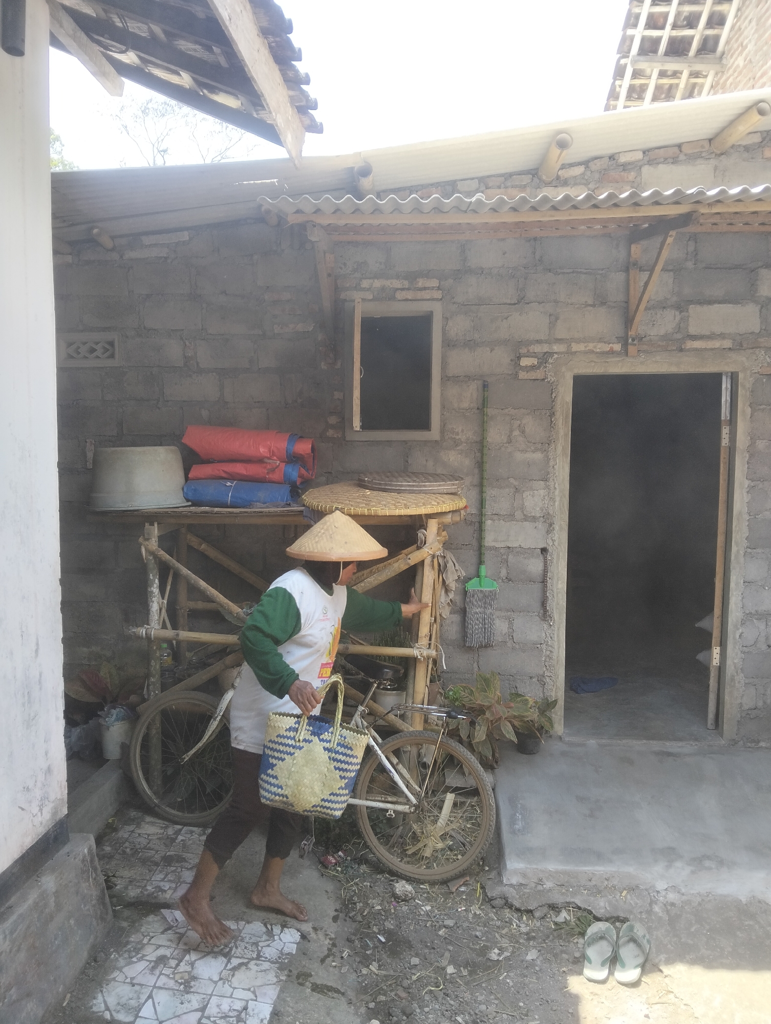 A person wearing a conical straw hat and a green-sleeved white shirt stands barefoot in front of a building. The person is carrying a woven basket with blue and yellow detailing and is holding onto the handlebars of a bicycle parked next to them. The building appears to be constructed of cinder blocks with a corrugated metal roof supported by bamboo. A wooden shelf unit with a large grey basin and two rolled-up bundles (one red, one blue) is visible behind the person. A green-handled mop hangs on the wall to the right of the shelf. The ground in front of the building is uneven and partially paved with cracked tiles. A doorway with a dark interior is to the right of the mop. Two grey flip-flops are placed on the ground near the doorway. The scene suggests an outdoor area in a rural or semi-rural setting, possibly a residential yard in Sumber Agung, Indonesia.