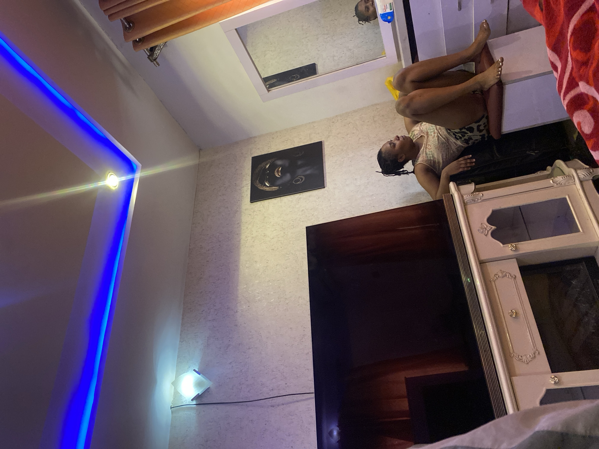 A woman sits on a chair in a bedroom in Giginyu, Nigeria, looking to the right of the frame. She has dark skin and braided hair, and is wearing a sleeveless top and shorts. The room has textured walls and a white entertainment unit beneath a large television. Above the television, a framed picture of a woman's face with gold accents hangs on the wall. To the right of the picture, a mirror reflects the woman and part of the room. The ceiling features blue LED strip lighting and a spotlight. Orange curtains hang on the right side of the room.