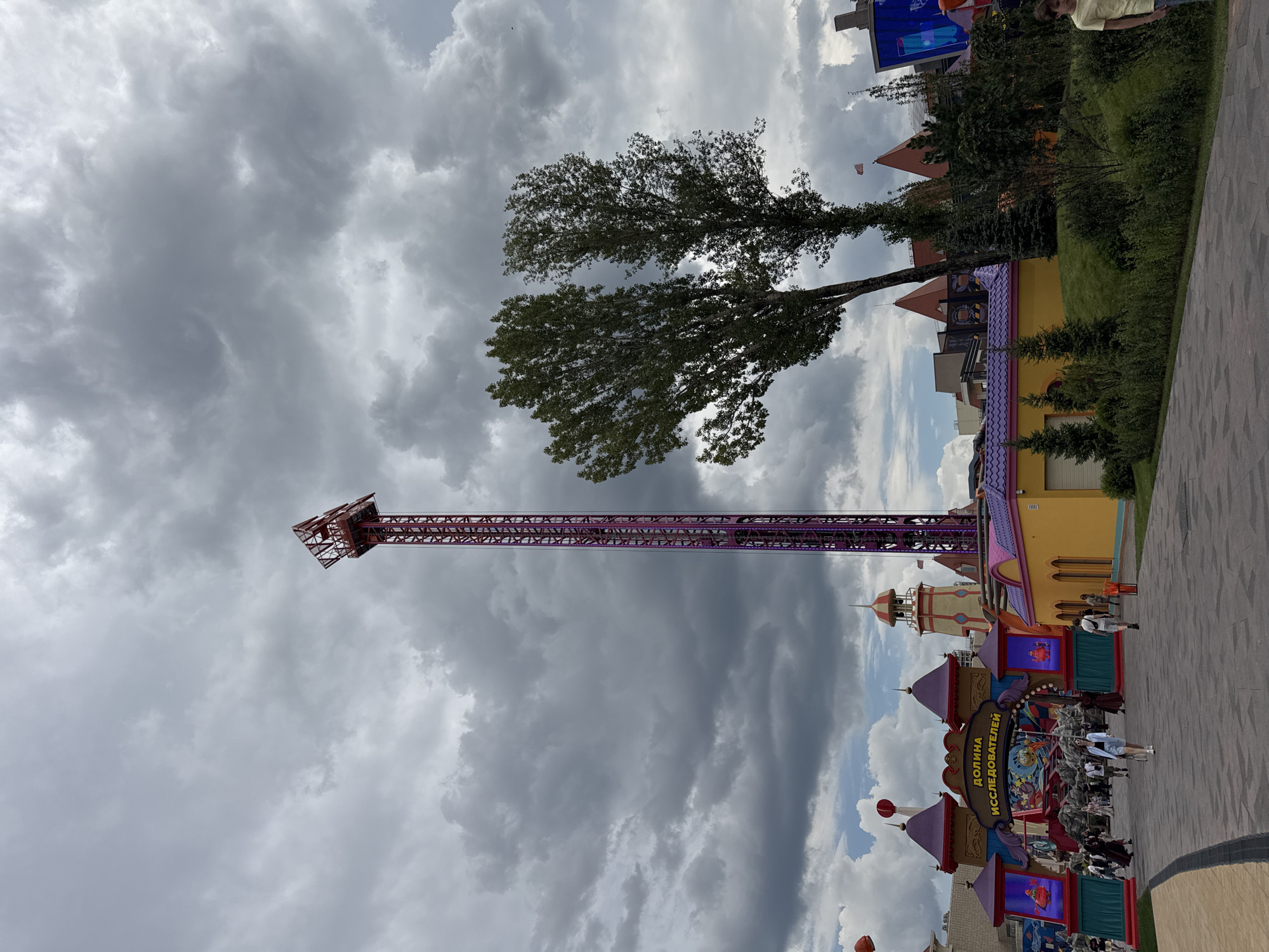 The image depicts an outdoor scene at an amusement park. A tall, purple and red thrill ride tower dominates the center of the frame against a cloudy sky. To the left of the tower is an elaborate amusement park entrance with colorful buildings, signage in Russian, and several people walking. Green trees and other park structures are visible. The location is Ostrov Mechty (Dream Island) in Moscow, Russia.