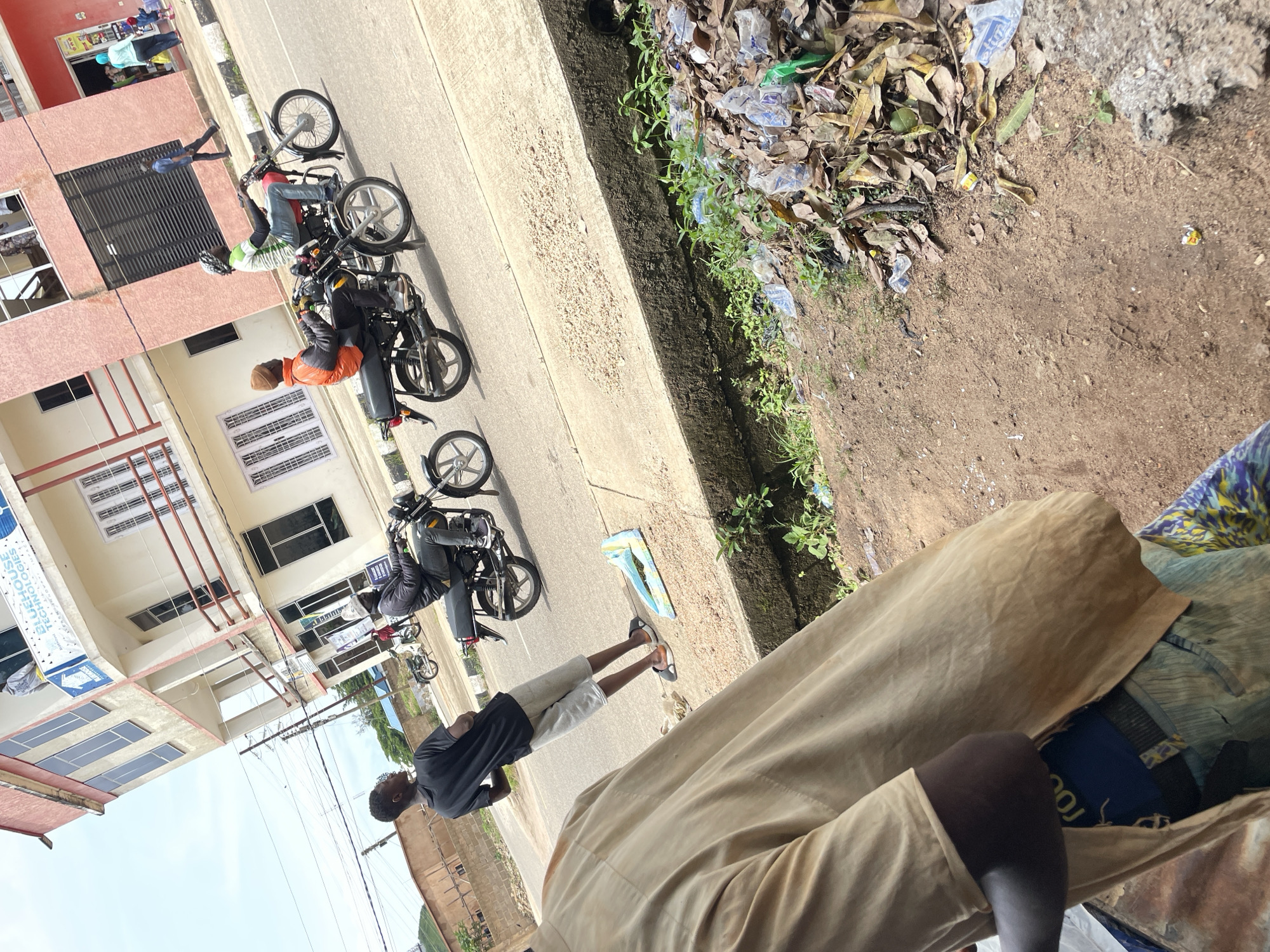 The image depicts a street scene in Jos, Nigeria, characterized by a busy road in front of a multi-story building. The building, with its light pink and white facade, features multiple windows and balconies, and bears signage indicating "BLUEHOUSE TECHNOLOGIES." Several individuals are present in the scene. In the foreground, a person wearing a tan garment is partially visible, with their back to the camera. Further down the road, a young man in a black t-shirt and grey shorts stands facing away from the camera. Dominating the mid-ground are two motorcycles in motion. On the left, a person in an orange jacket and brown beanie rides a black motorcycle. To their right, another person in a green and white striped shirt and blue jeans rides a red motorcycle. The road is paved, with a concrete barrier bordering a drain filled with leaves and plastic waste on the right side of the image. Other buildings and trees are visible in the background, suggesting an urban or semi-urban environment. The sky is overcast.