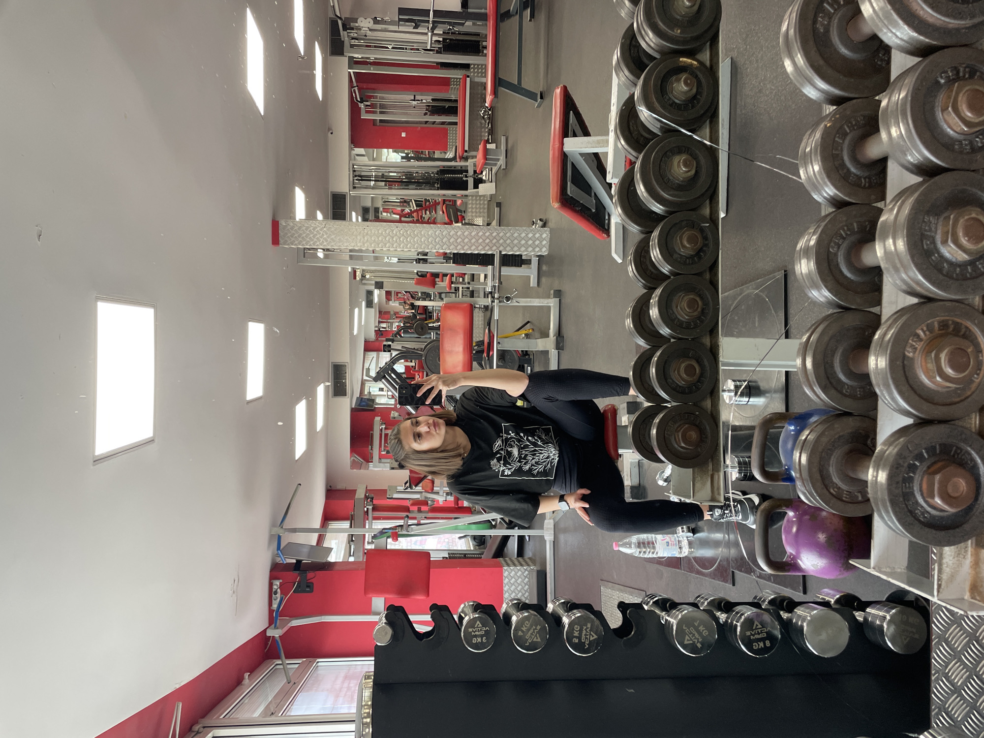A gym interior is photographed, featuring a young woman positioned to the left of the frame, apparently taking a selfie with her phone. She is dressed in black workout attire. Rows of dumbbells of different sizes and weights are visible in the foreground and along the right side, next to kettle bells and a water bottle. Weight training machines and equipment are located in the background. The location is likely in Sofia, Bulgaria.