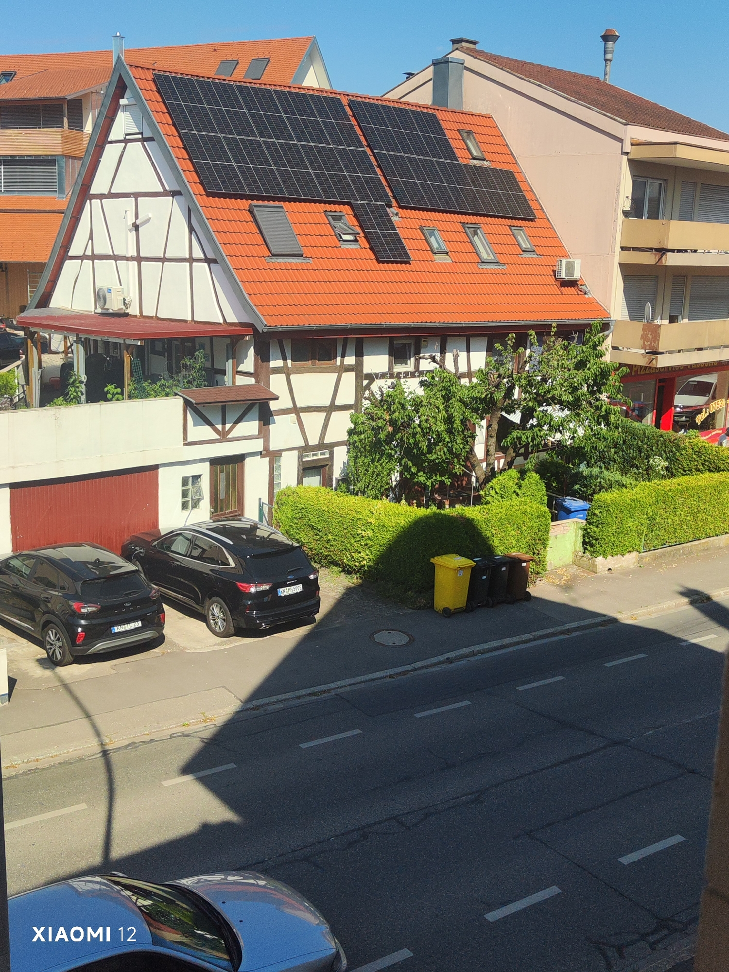 This media file depicts a sunny day in Constance, Germany. The scene shows a traditional German half-timbered house with a red-tiled roof and solar panels installed on the upper section of the roof. Next to it is a light pink building with balconies. In front of the half-timbered house, there is a red garage door, and two black SUVs are parked on a driveway. A neatly trimmed green hedge runs along the front of the property, and beyond that, a street with a blue recycling bin and several trash cans, including a yellow one, are visible. Lush green trees and bushes are also present. The time of day appears to be late morning or early afternoon, judging by the bright sunlight and the shadows cast by the buildings and trees. The clear blue sky indicates fair weather. The presence of the traditional architecture, the German license plates on the cars (KN-TL 23 and KN-MH 399E), and the city name "Constance" in the provided context pinpoint the location to Constance, Germany. The image is watermarked with "XIAOMI 12" in the lower left corner. There is no activity or event taking place; it is a static scene of a residential area.