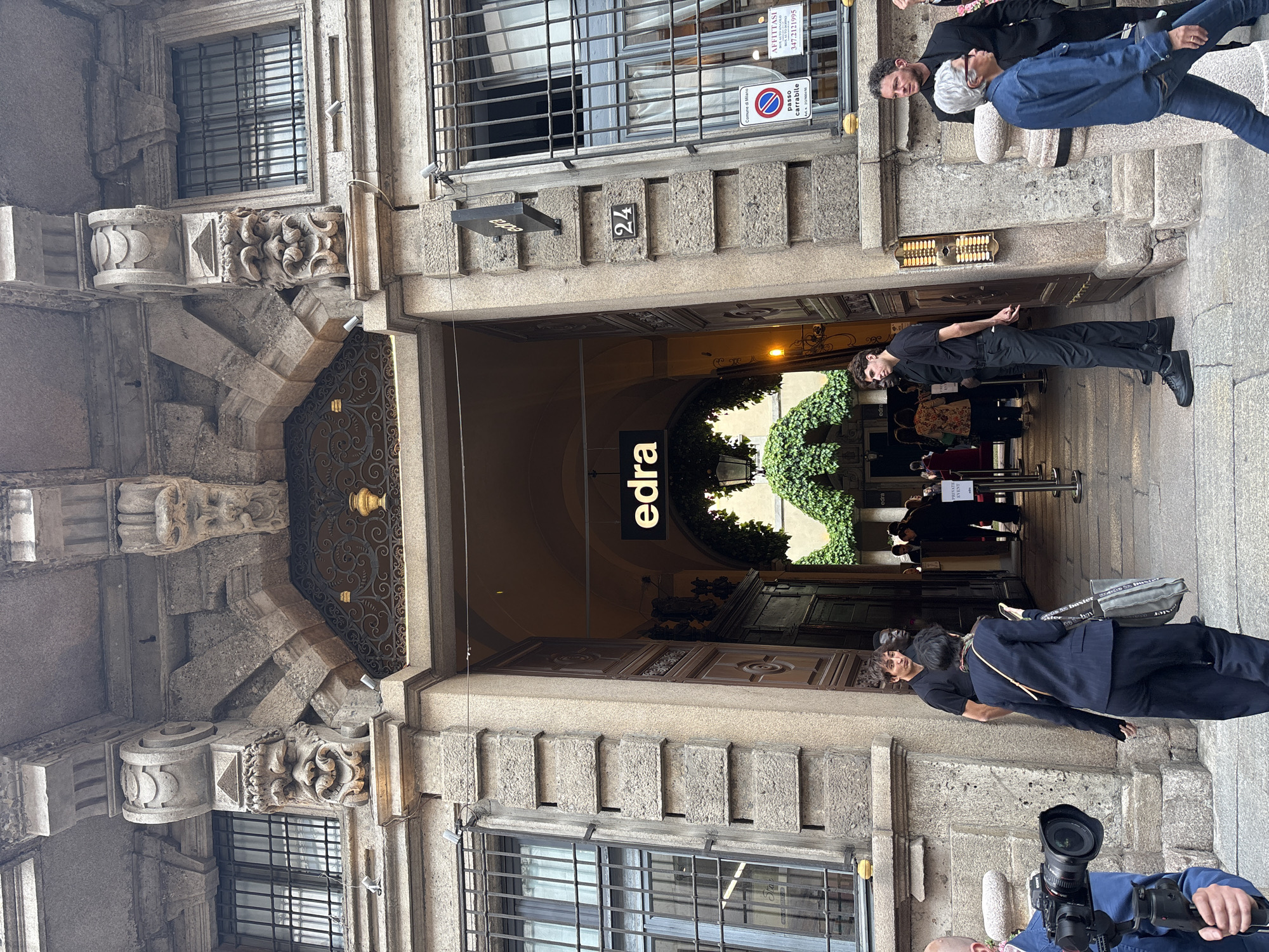 The image depicts a daytime scene outside a historic, ornate stone building in Milan, Italy. The central focus is a large arched entrance leading into an interior space, possibly a courtyard or an event venue. Above the arch, a prominent black sign displays the word "edra," and the building number "24" is affixed to the facade.
Several people are visible in and around the entrance. A man in a black shirt and pants stands upright near the archway, with another man in a blue jacket and jeans beside him. Further to the right, two men lean against a stone balustrade. In the foreground, a person is partially visible holding a professional camera with a large lens, suggesting a photographic or videographic activity. Inside the arched entryway, more people are gathered, and red barrier stanchions are visible, along with a partially obscured sign reading "PRIVATE EVENTS," indicating an organized gathering or exclusive access. Lush green foliage decorates the interior space beyond the arch.
Notable details include intricate stone carvings on the building's facade and robust window bars. On an upper window, a red and white "passo carrabile" (no parking/driveway) sign is displayed, along with a larger "AFFITTASI" (for rent) sign advertising "BOX AUTO MINGOLO BOX AUTO IKIPPIO" and a contact number "347.2121995." The lighting is natural and even, suggesting either an overcast day or a well-shaded street. The architectural style points to a typical European urban setting.