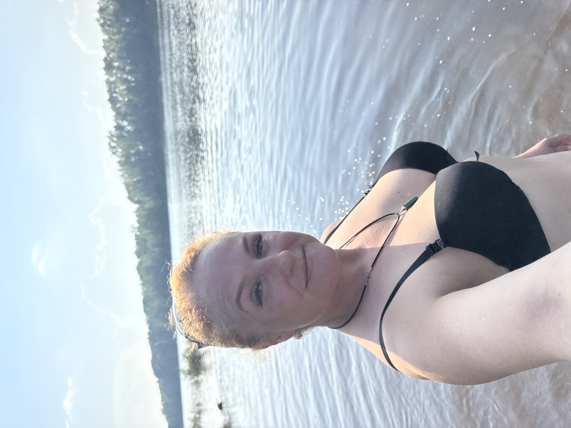 A woman with auburn hair is standing in shallow water, likely a river or lake, with a forested shoreline in the background under a clear sky with some clouds. She is wearing a black bikini and a necklace with a green pendant. The sun is shining, creating reflections on the water's surface. The image appears to be a selfie taken at waist level. The background details suggest a natural outdoor setting. The location is identified as Chaikovsky, Russia.