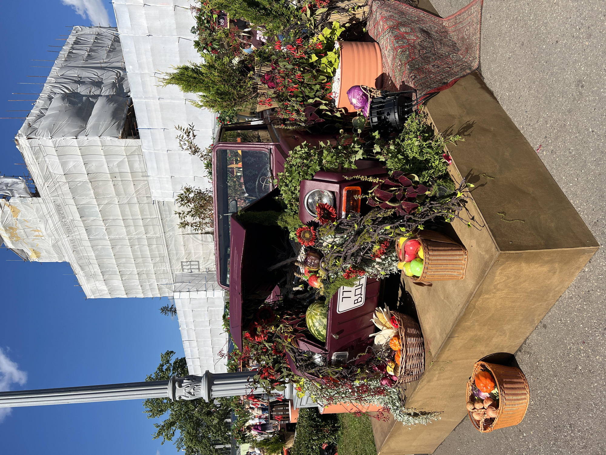 An outdoor still life display is set up on a raised wooden platform in front of a large building covered in white scaffolding and tarps, possibly under construction or renovation. A vintage maroon car, with its hood open, is the centerpiece of the display. The car is adorned with a profusion of plants, flowers, and autumn harvest produce, including a watermelon, pumpkins, apples, corn, and other vegetables. Woven baskets filled with similar produce are placed around the car and on the platform. A Persian rug is draped over the right side of the platform, and a spotlight is visible on the ground near the car. In the background, people are visible walking and standing near trees, suggesting a public space. The scene appears to be a festive or decorative arrangement, likely for an event or market. The building in the background has text visible on the scaffolding, and the license plate on the car displays Cyrillic lettering, suggesting a location in Russia. Given the architectural style and the context of such displays, it is plausible this scene is located in Moscow, Russia.