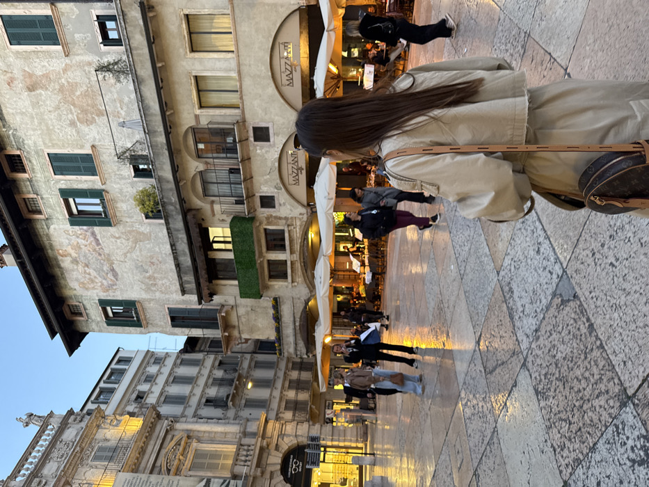 The image depicts an evening scene in Piazza delle Erbe, Verona, Italy. The square's surface is paved with large, irregularly shaped stone slabs, exhibiting a reflective sheen consistent with dampness.
In the foreground, a person is viewed from behind, with long, dark brown hair. They are wearing a light beige cropped jacket over a longer, similarly colored garment. A round brown Louis Vuitton shoulder bag with a dark brown monogram pattern hangs by a thin leather strap from their right shoulder. This individual is facing away from the camera, observing the scene.
The midground and background feature multiple historic buildings, illuminated by artificial light against a twilight sky. To the right, a multi-story building with an aged, light-colored stucco facade is prominent. It displays a faded fresco or mural on an upper section and numerous windows fitted with dark green wooden shutters, some open, some closed. Balconies with railings are present on various levels. The ground floor houses "Mazzanti Caffè," identifiable by its signage, with an extensive outdoor dining area featuring multiple tables, chairs, and large white market umbrellas, all brightly lit with warm, artificial light.
To the left, a more ornate, classical-style building with architectural details, columns, and sculptures on its roofline is visible. Its ground floor hosts a "LUXURY EYEWEAR OUTLET," illuminated with bright lights. Further back on the left, another multi-story building with a simpler design and numerous windows completes the urban backdrop.
Approximately 15-20 individuals are scattered throughout the square and the outdoor café area. Some are walking across the paved surface, while others are seated at the café tables, engaged in dining or conversation. On the left midground, a group of three individuals includes one wearing light blue jeans and a brown top, and another in a dark suit holding a white bag. The overall scene suggests an active public square during the early evening hours.