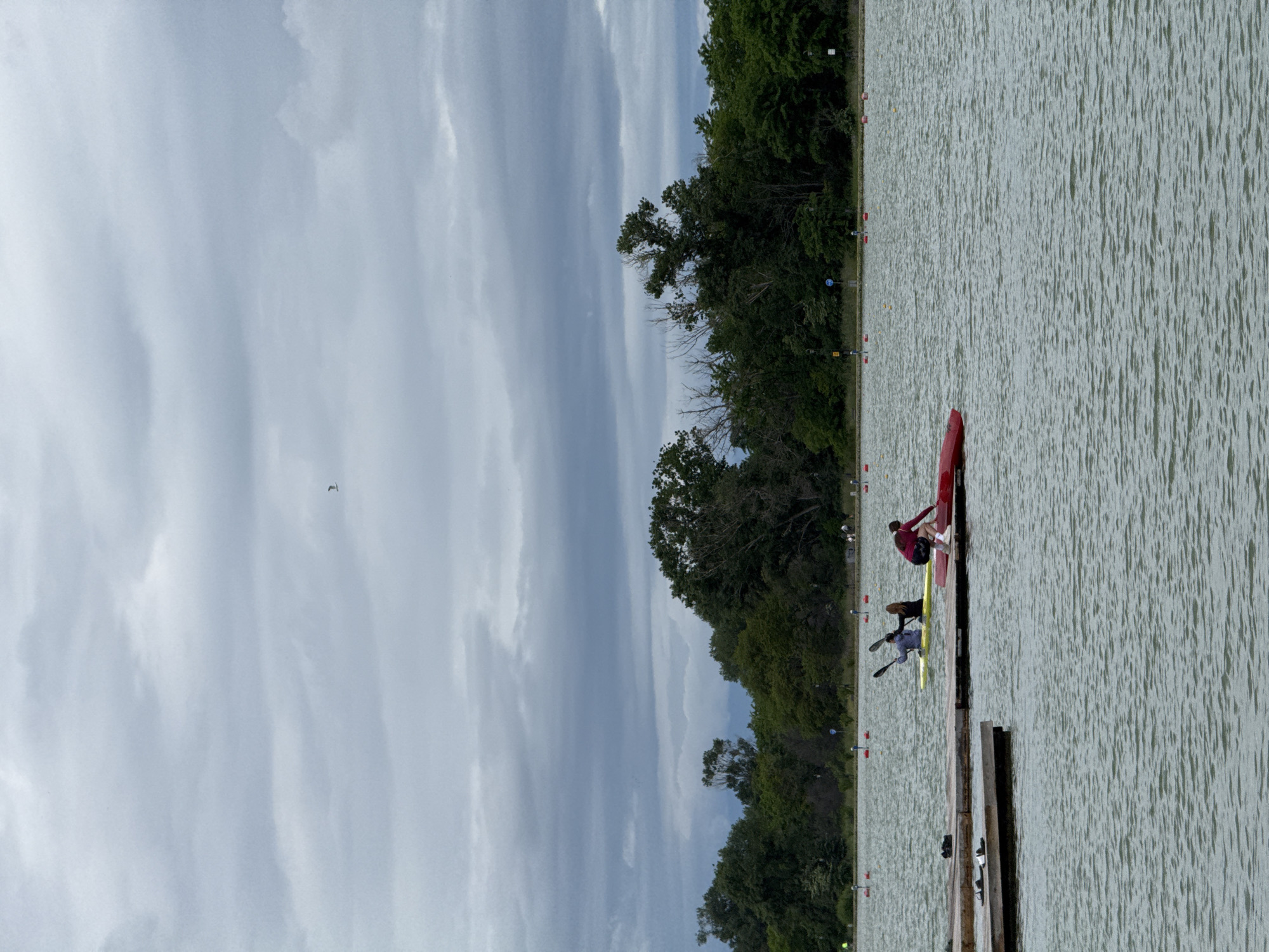 The image depicts a rowing scene in Plovdiv, Bulgaria, presumably on the Plovdiv Rowing Canal. In the foreground are two racing kayaks docked at a wooden pier. One kayak is red, the other is yellow. There are two people in the kayaks, positioned to begin rowing. A third person in a burgundy shirt is standing on the pier, possibly assisting. The water is slightly rippled, suggesting a mild breeze. The background is a line of dense green trees along the water's edge, marking the edge of the canal. The sky is overcast with a striated cloud formation. Racing markers are visible lining the course.