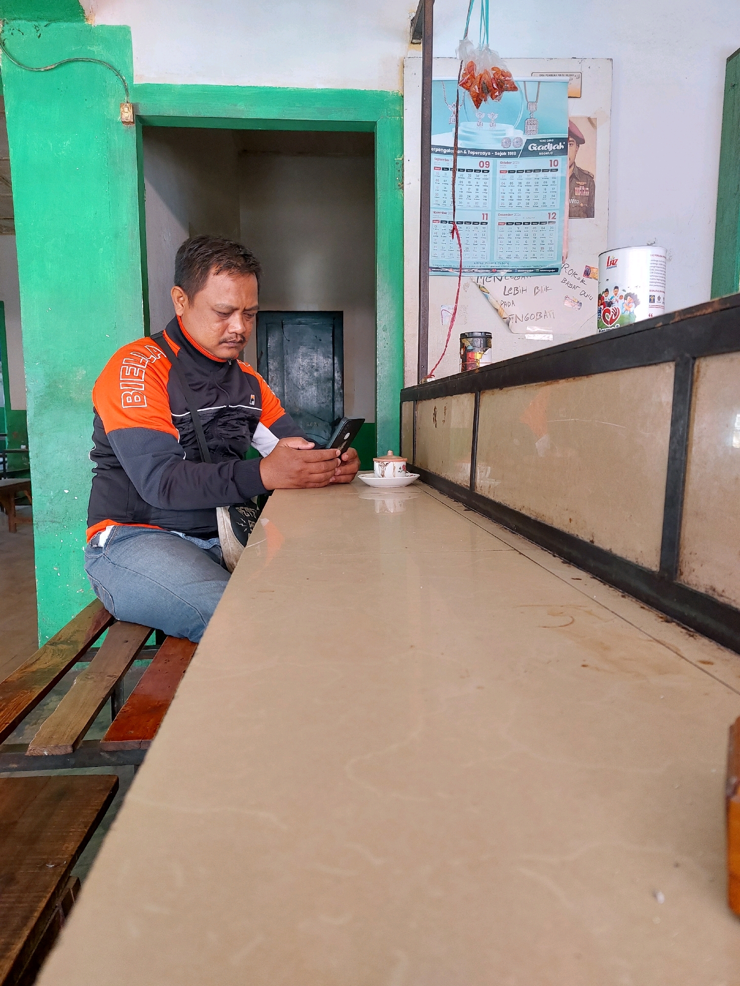 In Sumuragung, Indonesia, a man is seated on a wooden bench at a counter, engrossed in his smartphone. He is wearing a black and orange jacket over a white shirt and blue jeans. A small cup of coffee sits on a saucer to his right on the counter. The counter itself appears to be made of a beige, speckled material with a dark metal frame. Behind the counter, a wall displays a calendar featuring jewelry and a picture of a person in uniform. Various items, including a tin can and a tube, are visible on shelves behind the counter. The surrounding environment features a green-painted door frame and wall, with exposed wiring. Wooden slats form the seating area for the bench.
