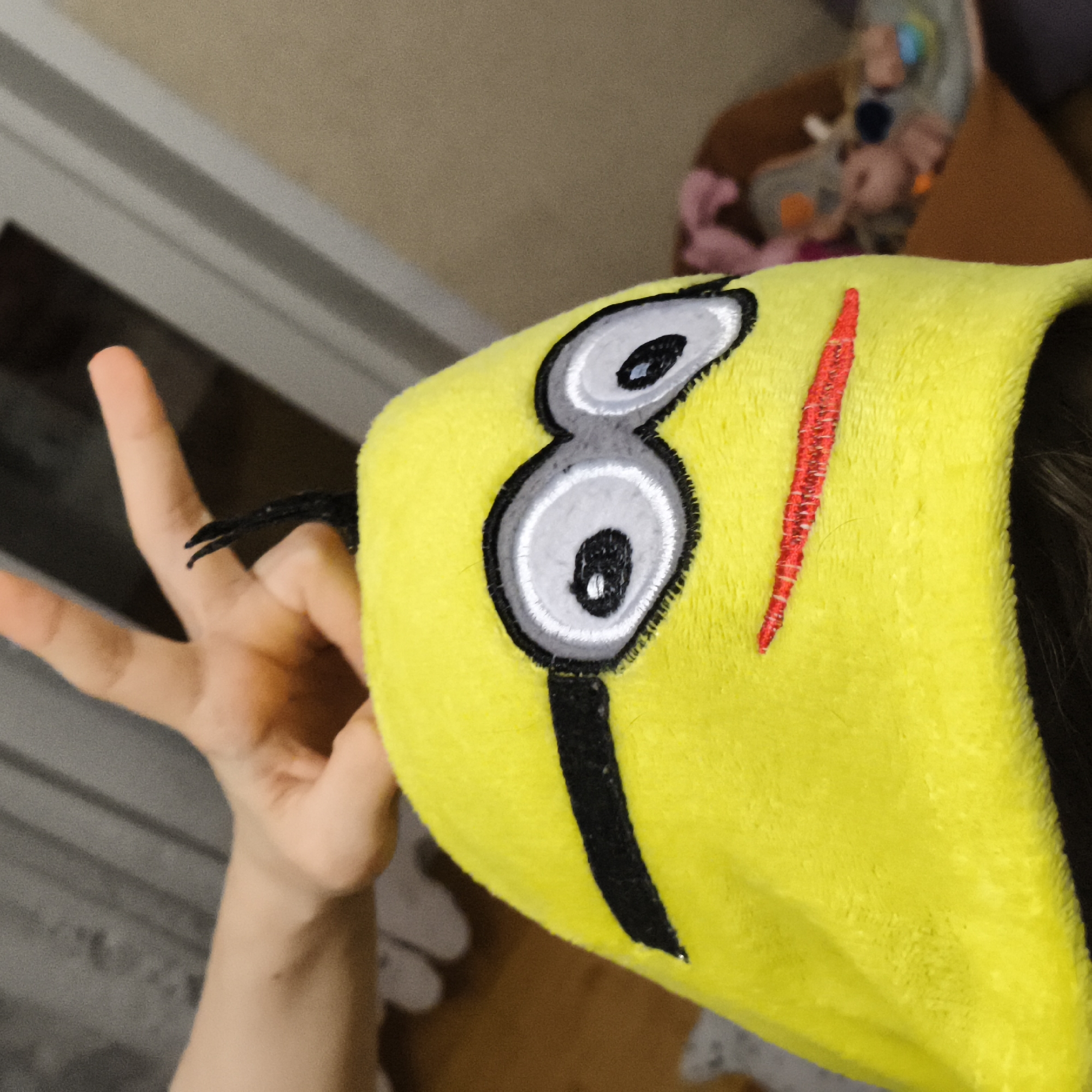 The image shows a person wearing a yellow minion-themed hood, extending two fingers in a "peace" sign. The hood features embroidered details of the minion's eye and goggle design. The background appears to be an indoor residential setting. There is no clear information indicating the location to be Chaikovsky, Russia.