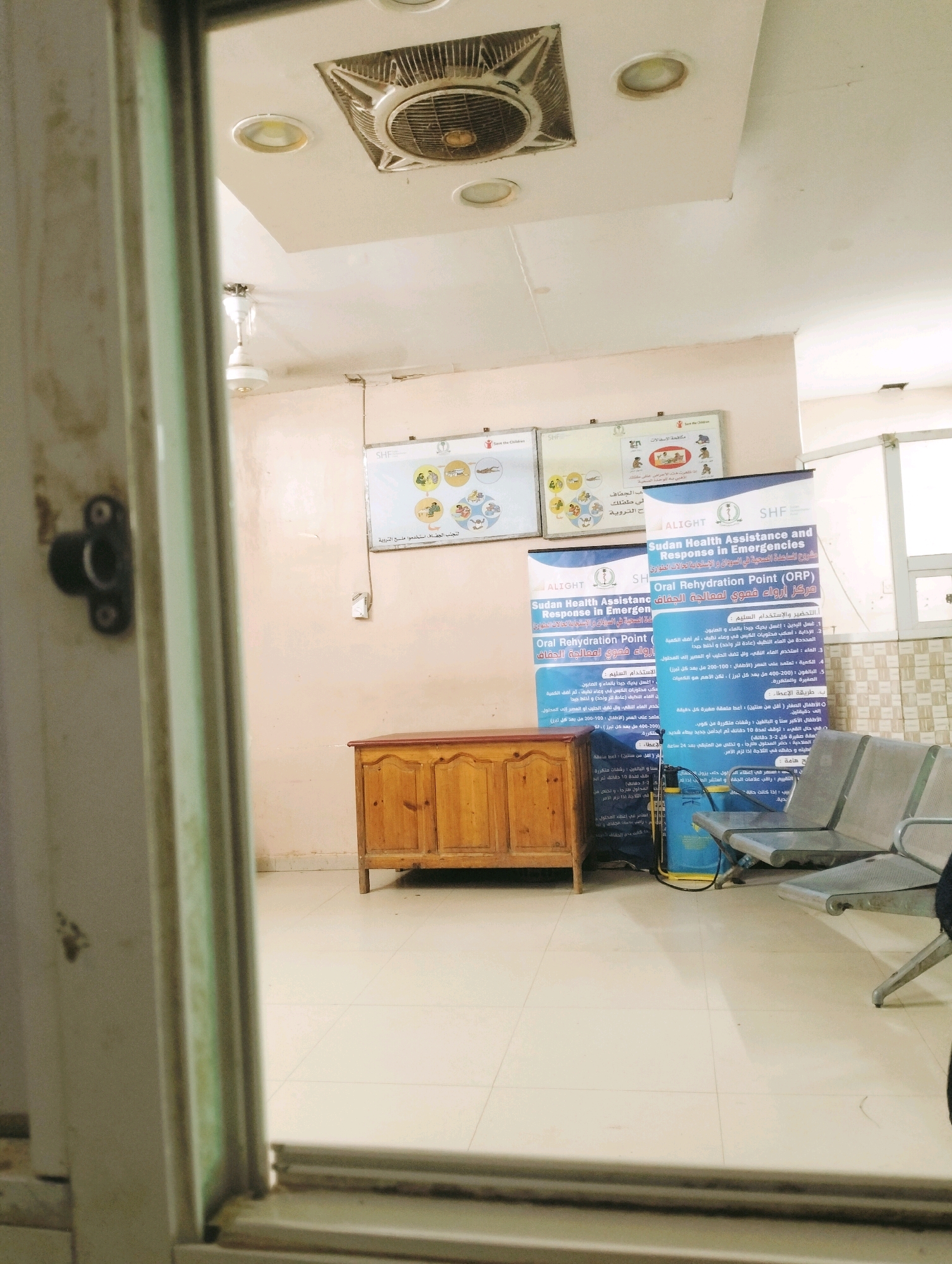 The image depicts an indoor waiting area, possibly a clinic or health facility, within the city of Al Gadaref District, Sudan. The scene is viewed through a doorway or opening, with the foreground occupied by the frame of the opening. The room itself is brightly lit and appears to be sparsely furnished.
Dominating the background are two large posters on the wall. The poster on the right, in particular, is a large blue banner with white text, which appears to be educational material related to "Sudan Health Assistance and Response in Emergencies" and "Oral Rehydration Point." To the left of this poster, another smaller poster is visible.
In the center of the room, a wooden cabinet with two doors stands on the floor. To the right of the cabinet, a set of metal chairs, arranged in a row, are visible. A blue plastic container, possibly a water tank or a cleaning equipment, is situated between the cabinet and the chairs.
The ceiling features a square fan and several circular lights. The walls are painted a light, neutral color. The floor is tiled with large, square, light-colored tiles. The overall impression is that of a functional, but perhaps not recently updated, public service space. No individuals are clearly visible in the image, suggesting the area may be momentarily unoccupied or the photo was taken from a discreet vantage point.
