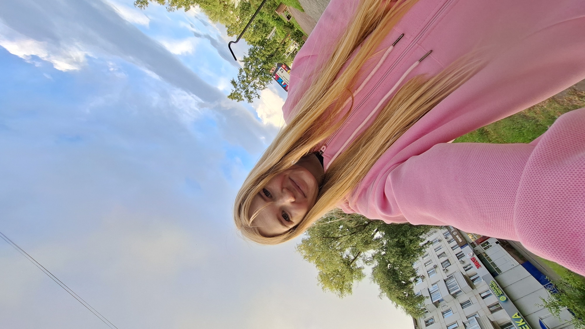 An outdoor, waist-up, selfie-style photograph features a young adult female with long, straight blonde hair and a smiling expression. She is wearing a pink hooded jacket. The background includes a residential building with multiple floors and balconies, trees, a paved walkway, and a cloudy sky. A sign with Russian text is partially visible on the building. The lighting suggests it is daytime, possibly late afternoon or early evening. The overall environment appears to be a residential area in the city of Chaikovsky, Russia.
