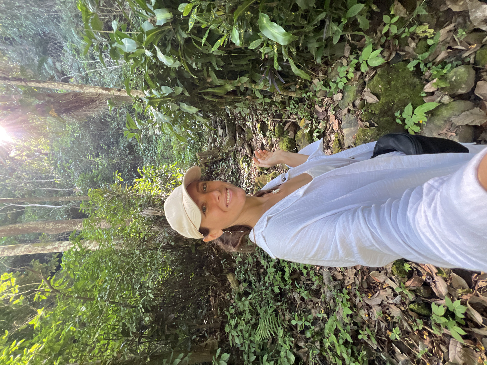 A woman wearing a white shirt and a beige baseball cap smiles at the camera while standing on a stone path in a lush, green forest. Sunlight streams through the trees, creating a dappled effect on the foliage. The path is lined with moss-covered rocks and dense vegetation, including ferns and large-leaved plants. The woman has a necklace with pearls. She is holding a black bag. The scene appears to be a nature trail, possibly in a tropical or subtropical region. There is no indication of the city of Busungbiu, Indonesia, within the image.