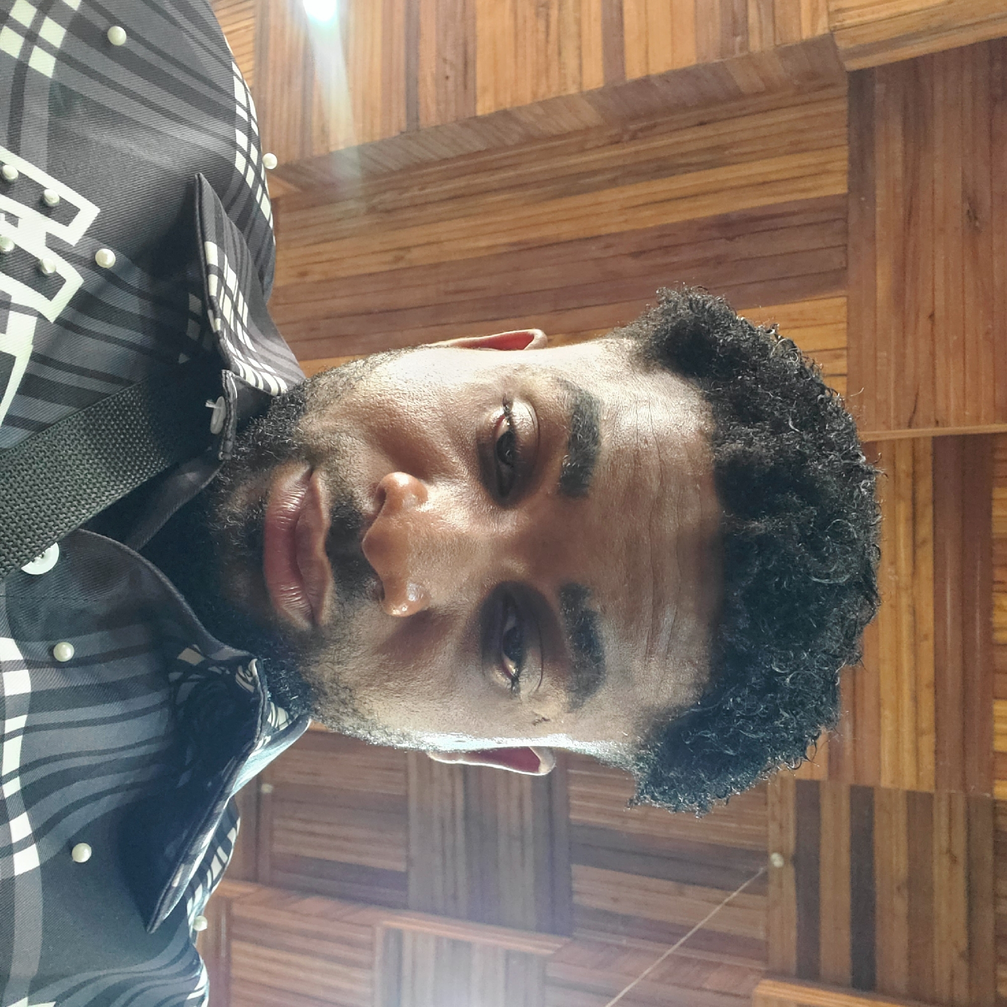A man with dark curly hair and a beard is posing in front of a wooden wall. He is wearing a black and white plaid shirt with pearl-like embellishments. The scene appears to be indoors, the location is the city of Nkpor, Nigeria.