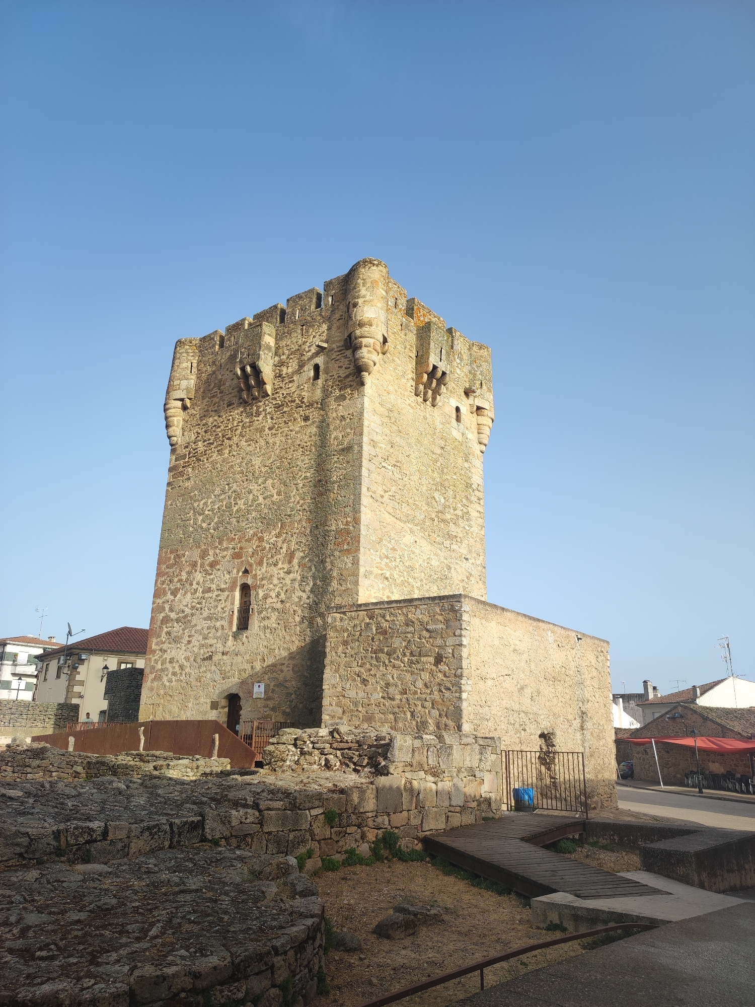 A wide, low-angle, exterior, daytime shot captures the ancient stone Tower of Sobradillo, located in the municipality of Sobradillo, Spain. The tower, constructed from rough-hewn, tan and reddish-brown stones, dominates the frame. Its battlements and weathered facade suggest a long history. To the right of the tower, a smaller, lower stone structure is attached. In the foreground, the remnants of ancient stone walls and rubble are visible, indicating an archaeological site or ruins. A wooden walkway with a handrail leads towards the main tower. A metal gate, behind which a blue object is partially visible, is situated on the right side of the walkway. Beyond the tower, to the left, are modern buildings with tiled roofs and white facades. To the right, street elements are visible, including a street, a red awning over what appears to be an outdoor seating area with tables and chairs, and a parked car. The sky above is a clear, bright blue. There are no people visible in the image.