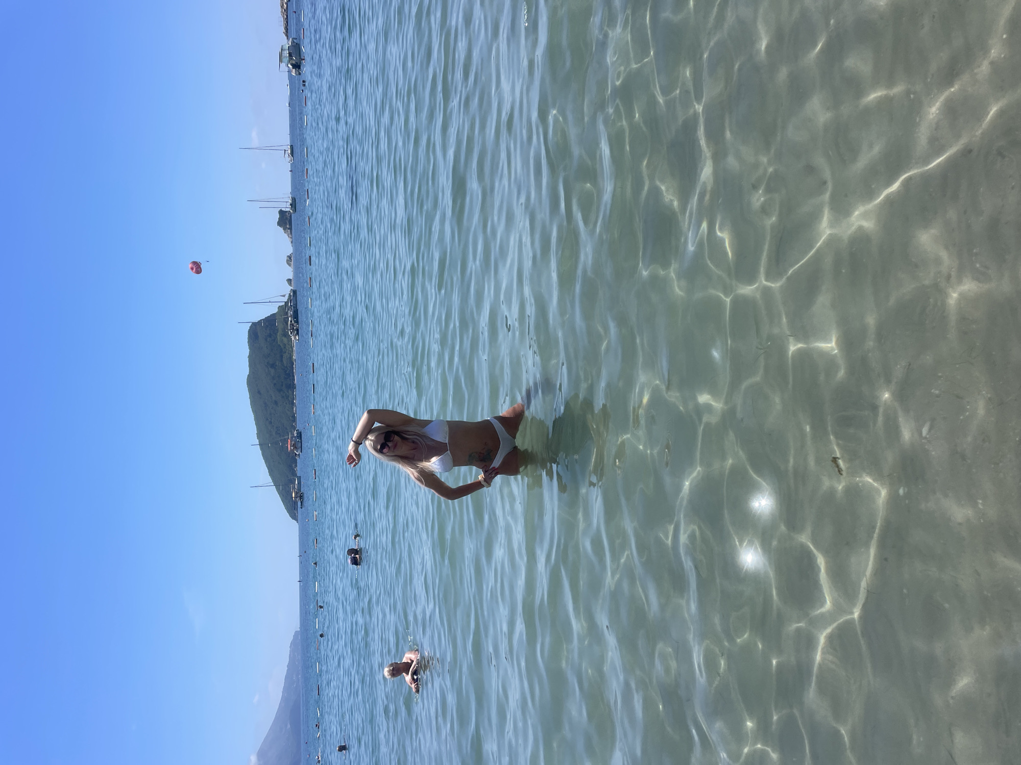 A woman in a white bikini stands in clear, shallow water with her hands on her hips and her arms raised above her head. She is wearing sunglasses and has blonde hair. The water is calm and reflects sunlight, creating shimmering patterns on the sandy bottom. In the background, other people are swimming and enjoying the water. Several boats are anchored offshore, and a green, tree-covered hill rises from the coastline. The sky is a clear blue, indicating a sunny day, likely in the late morning or early afternoon. A red parasail is visible in the distance, suggesting a tourist destination with water sports activities. The scene is set in a coastal area, possibly a beach or bay, with calm water and a backdrop of hills and boats.