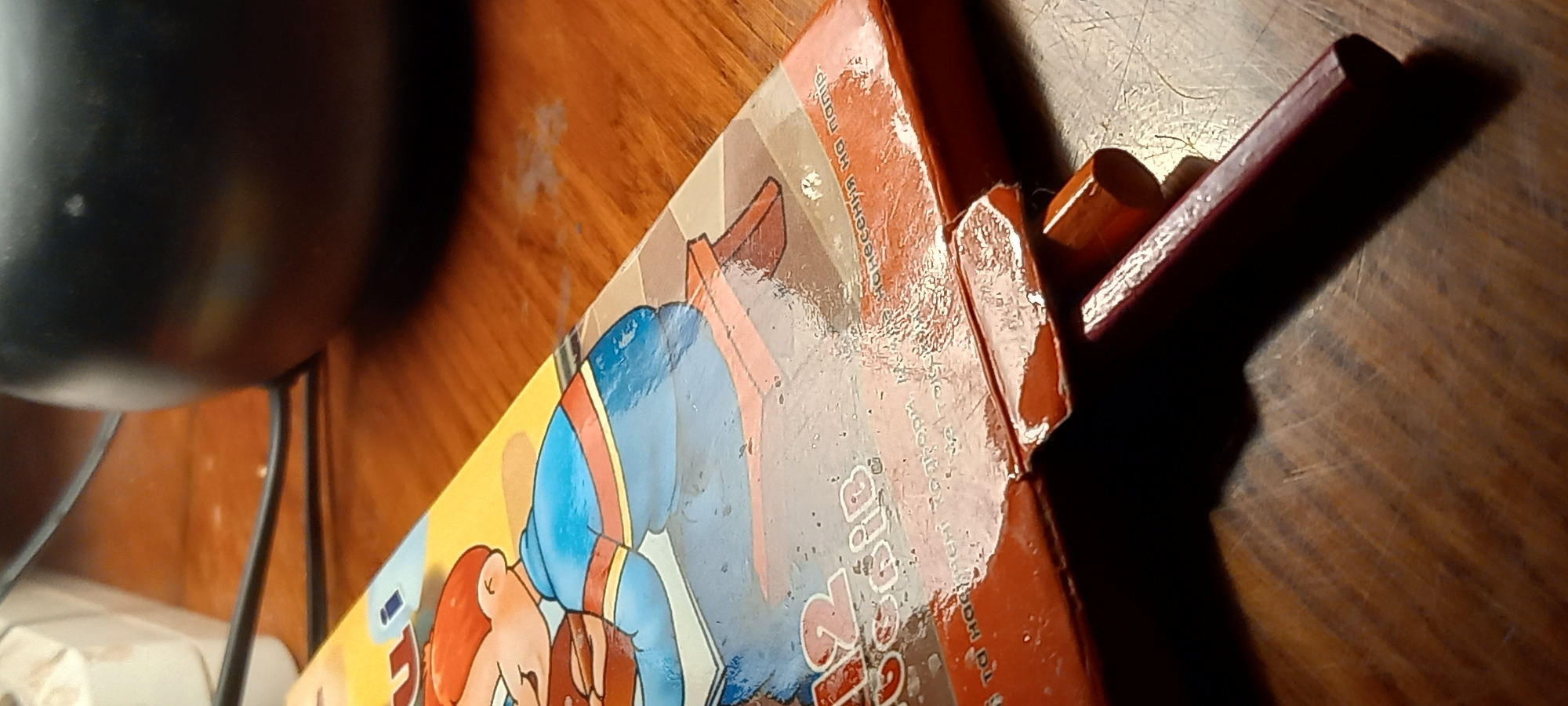 The image presents a close-up, slightly angled view of various objects arranged on a dark wooden surface, likely a table. The primary subject is a colorful, somewhat worn cardboard box, possibly for a game or art kit.
On the visible side of the box, there's a cartoon illustration of a character with red hair, wearing a blue top with orange/yellow stripes, and blue trousers. The character appears to be in an active pose, possibly running or jumping. The surface of the box shows signs of wear, including scuffs, scratches, and a somewhat shiny, possibly peeling, clear film or laminate, with some moisture or residue visible, particularly along the top edge.
Visible text on the box, primarily in Cyrillic, includes fragments like "...НОСЕННЯ НА ПАПІР" (likely part of "Нанесення на папір," meaning "Application on paper"), "...подорож" ("journey"), and parts of other words such as "Тоді Ма...". A prominent stylized text, possibly part of the title, reads "...ig Rip".
To the right of the box, two small, dark reddish-brown, rectangular objects are visible, resembling crayons, wooden blocks, or small tools. On the far left, a portion of a dark, curved, metallic object, possibly a pot or pan, is visible, along with a thin black wire or stand. A small white object rests in the lower-left corner.
The lighting appears to be indoor and indirect, creating some shadows on the wooden surface. The overall scene suggests an ordinary indoor setting, possibly a home, in Kryvyi Rih, Ukraine, given the provided context. No specific activity is taking place beyond the arrangement of these objects.