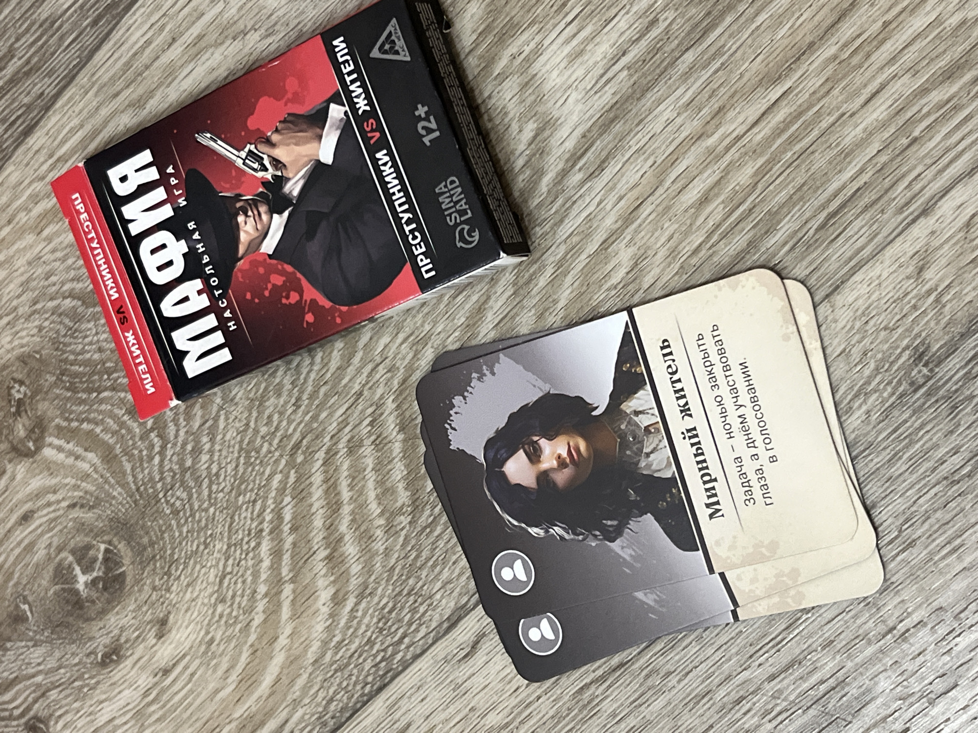 The image shows a tabletop view of a board game titled "Mafia" on a wooden surface. The game box, which is predominantly red and black, features an illustration of a man in a suit and hat holding a pistol. Text on the box indicates it is a "tabletop game" and "criminals vs. residents." The bottom of the box shows the brand "SIMALAND" and the age rating "12+". In the foreground, several cards are fanned out. One visible card depicts a woman with dark, curly hair and has the title "Mirnyy zhitel" (Peaceful resident) printed on it, followed by a description of a task. The background is a close-up of a wooden surface, likely a table, with visible grain patterns and knots. The lighting suggests the image was taken indoors with overhead light. There is no specific indication of the location within the city of Дачное, Russia.