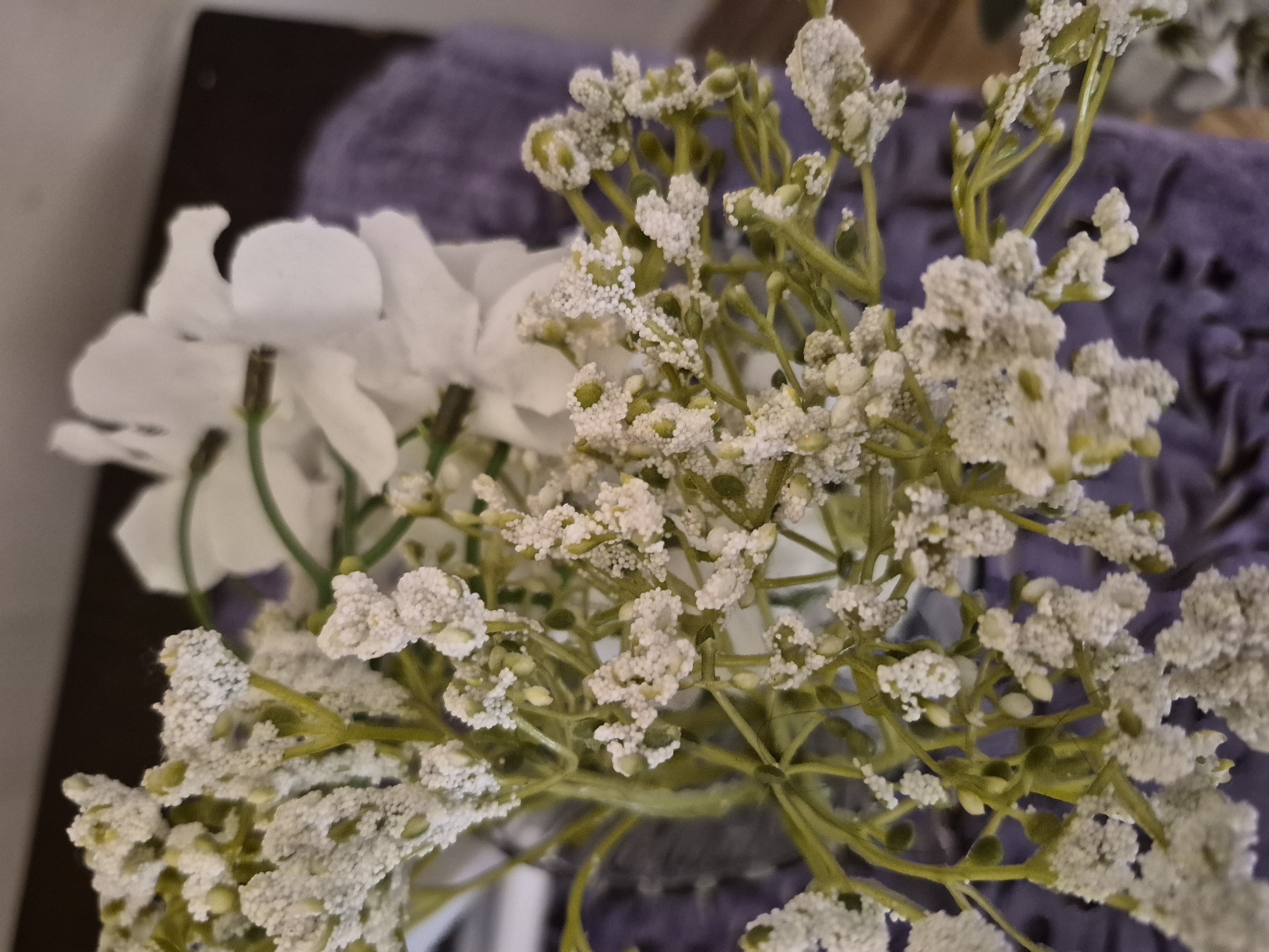 The image displays a high-angle, close-up view of an artificial floral arrangement. The foreground is dominated by clusters of small, white, textured flowers resembling baby's breath, connected by green artificial stems and immature buds. These small flowers exhibit a granular or beaded surface texture. In the upper-right background, several larger, smooth white artificial flowers are partially visible and softly out of focus. The background consists of a blurred purple textile material, possibly fabric or a towel, situated below the flowers, and a dark, indistinct surface (potentially a table or shelf) behind them. No individuals are present, and no actions or interactions are depicted. The specific location, including any city or country, is not discernible from the visual content provided.