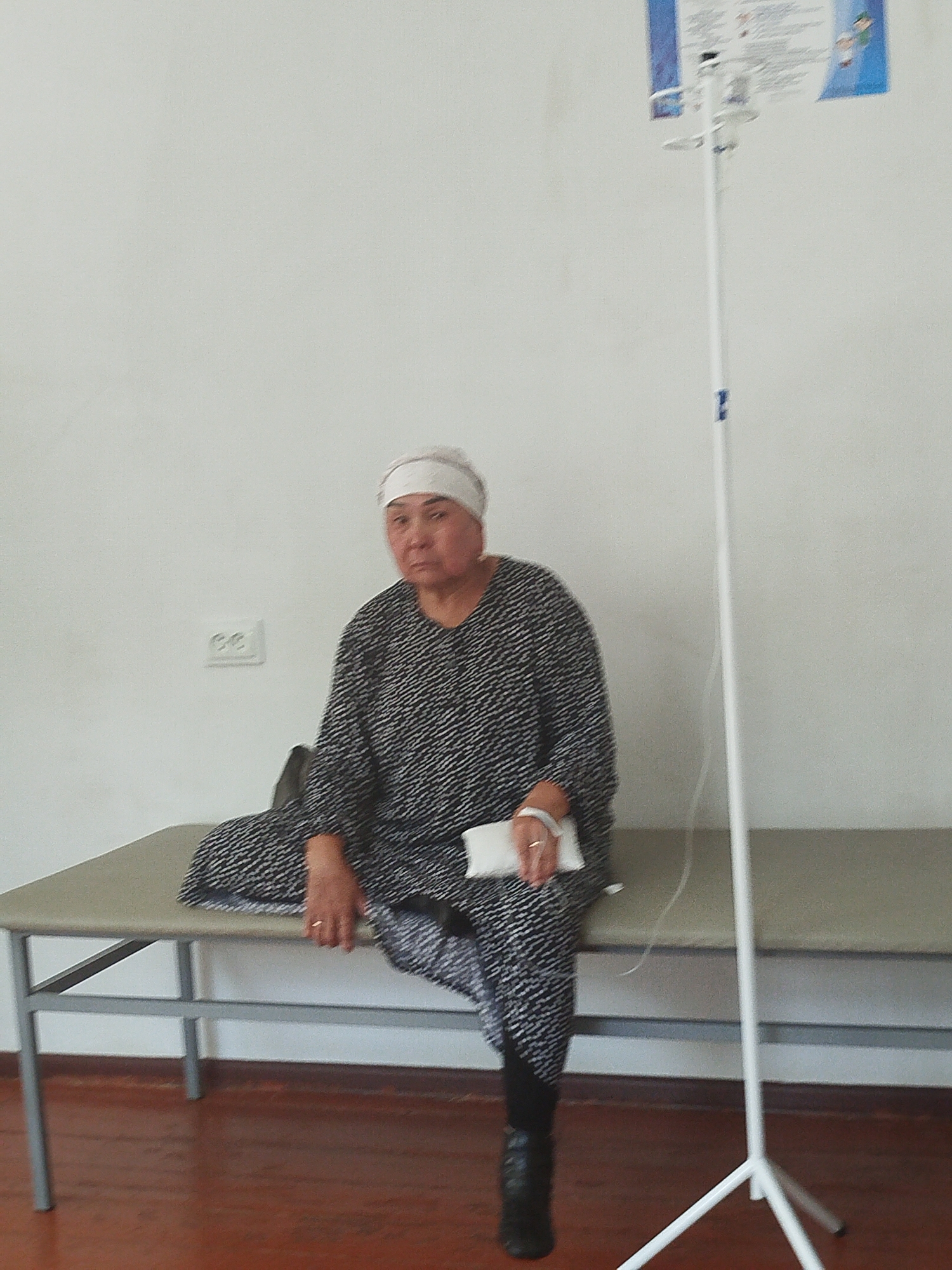 An elderly woman with a white headscarf is seated on a medical examination table in a room with white walls. She is wearing a black and white patterned dress and black boots. Her right hand is resting on a white packet, and she is wearing a ring on her ring finger. To her right, a white IV stand with a bag of fluid is visible, with tubing running down towards the floor. A double electrical outlet is mounted on the wall to the left of the woman. The examination table has a light-colored surface and a gray metal frame. The floor is made of dark red wood. The scene appears to be in a medical facility in Orozbekov, Kyrgyzstan.