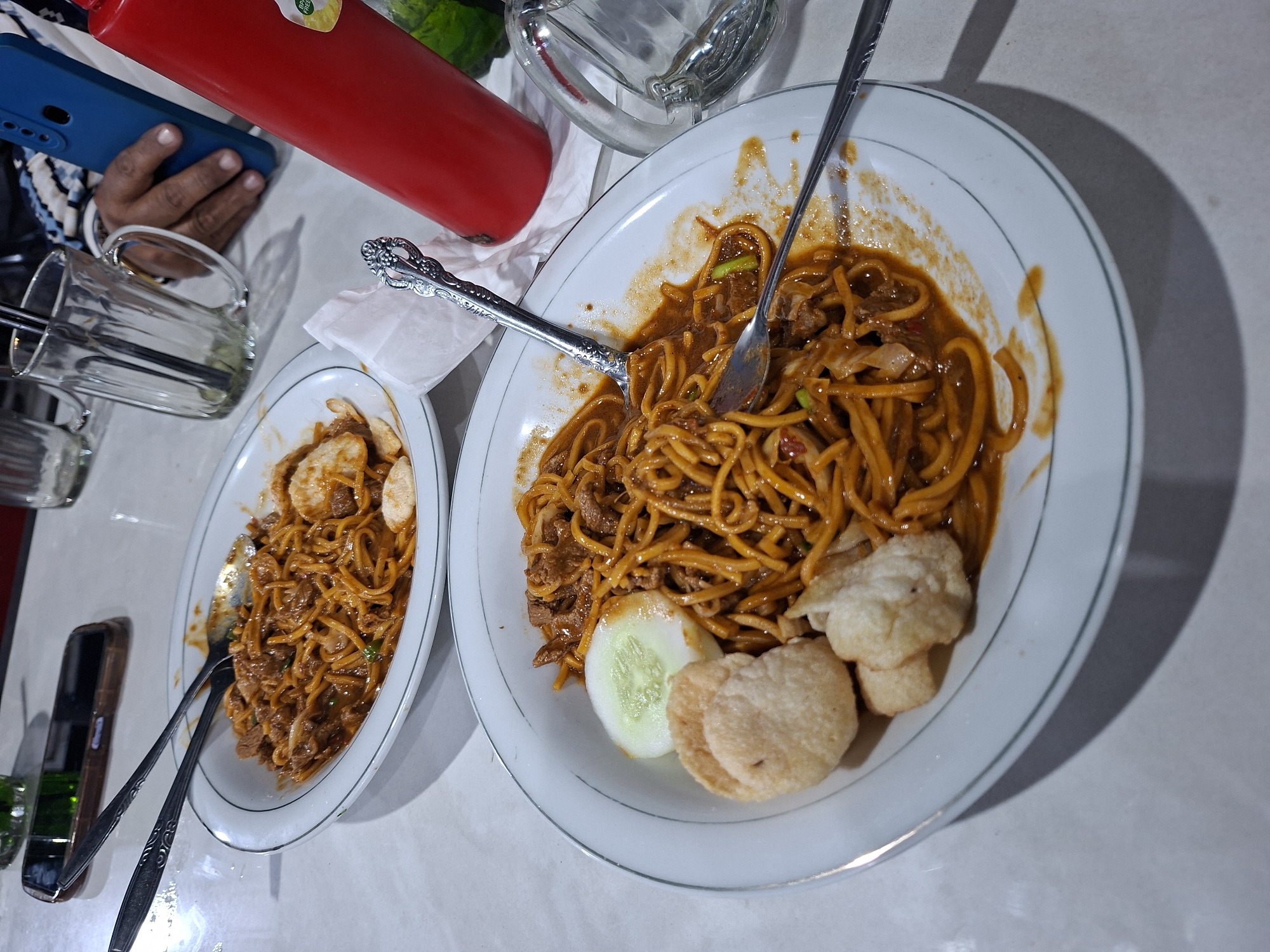 The image depicts two plates of Mie Aceh, a noodle dish, on a table. Mie Aceh consists of thick yellow noodles mixed with a dark brown sauce, meat, and vegetables. The larger plate also has cucumber slices and crackers. Two forks and spoons are present on the plates. A person is holding a blue smartphone in the upper left, and a red bottle is nearby. Glassware is visible. The location is Lhokseumawe, Indonesia.