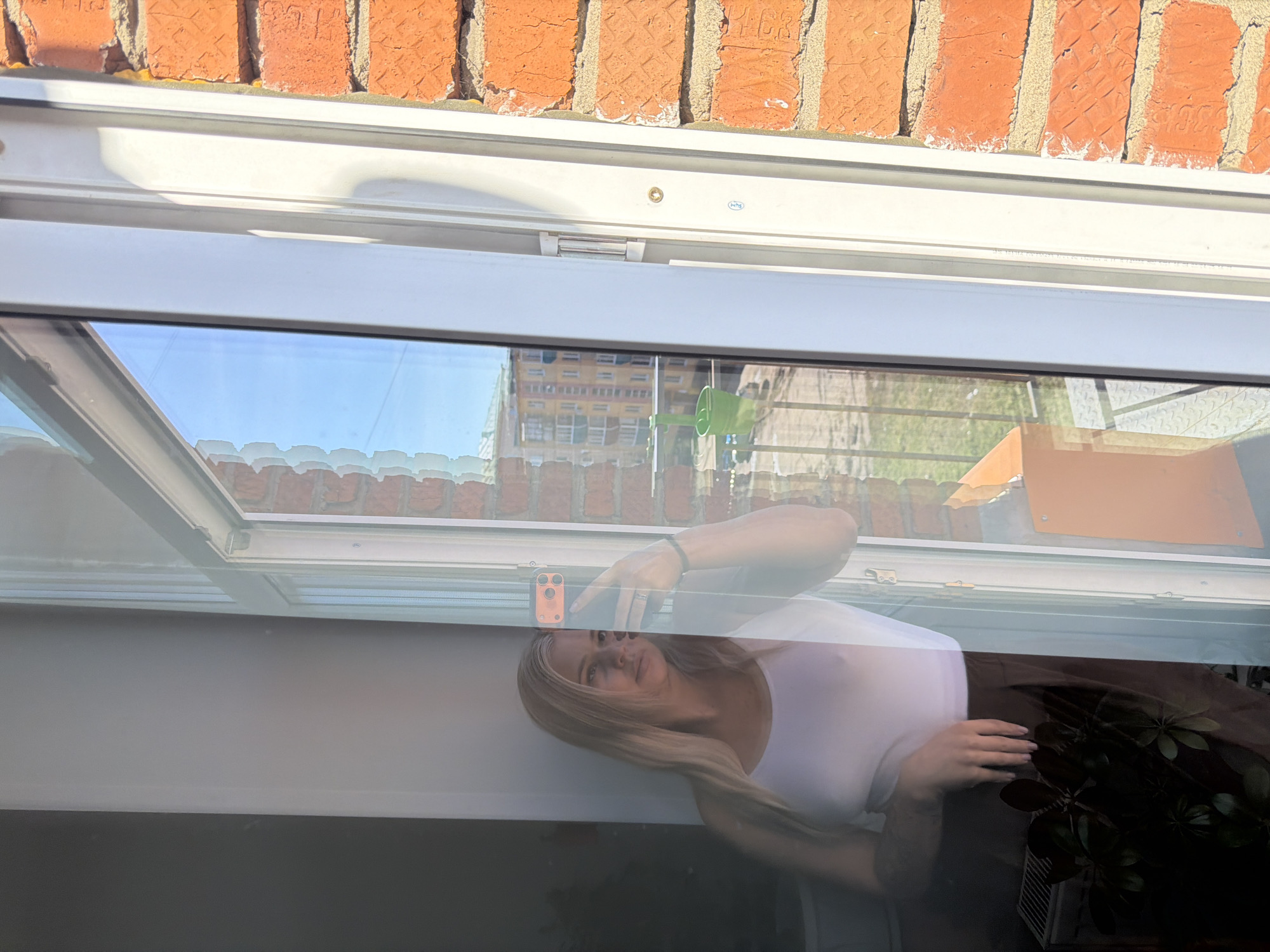 The image, rotated 90 degrees clockwise from a typical upright perspective, primarily captures reflections on a glass surface, likely a window or skylight, looking out onto an exterior scene. The dominant subject is the clear reflection of a young woman with shoulder-length blonde hair, wearing a white top. She is holding a smartphone with an orange case in her right hand, suggesting she is taking a photo or selfie. She has a ring on one finger and a dark bracelet on her wrist, and her expression is neutral with a slight smile.
To the lower right of her reflection, a dark-leaved houseplant is visible. Above the reflection, the frame of the window or skylight is prominently featured, made of white material. Beyond this frame, an exterior brick wall with orange-red bricks and light mortar is visible. Through the glass, in the background behind the reflection, a multi-story building with a yellow facade and numerous windows is faintly reflected, along with a bright blue sky, indicating clear, sunny daytime weather. A small green plastic item hangs near the reflected building.
On the white window frame, there is a small blue oval sticker with white text, which appears to be a manufacturer's label or identifier, possibly "HNS" or "PNS" followed by numbers. Other very faint, illegible text is also present on the frame. The strong natural light suggests it is daytime. The scene depicts a moment captured indoors through reflective glass, offering a view of an exterior urban or residential environment in Sarapul, Russia.