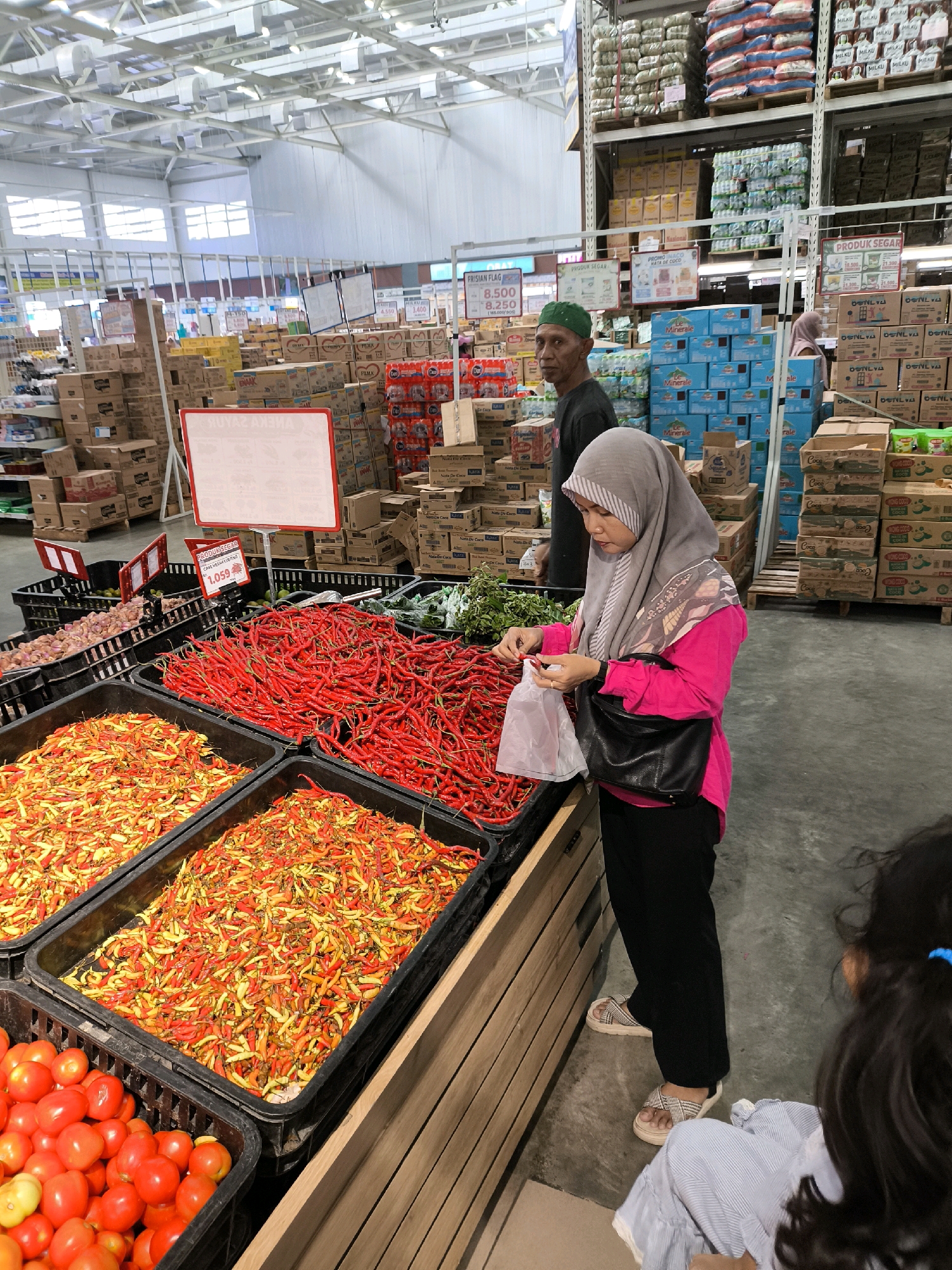 The image depicts the produce section of a supermarket in Gorontalo, Indonesia. A woman wearing a grey hijab and a bright pink shirt is standing in front of a display of fresh red chili peppers. She is holding a white plastic bag and appears to be selecting chilies. To her left, there are bins filled with various types of chili peppers and tomatoes. In the background, shelves stocked with goods and other shoppers can be seen. A man wearing a green cap stands behind the produce display, looking towards the camera. The environment suggests a bustling retail setting with ample lighting and organized merchandise.