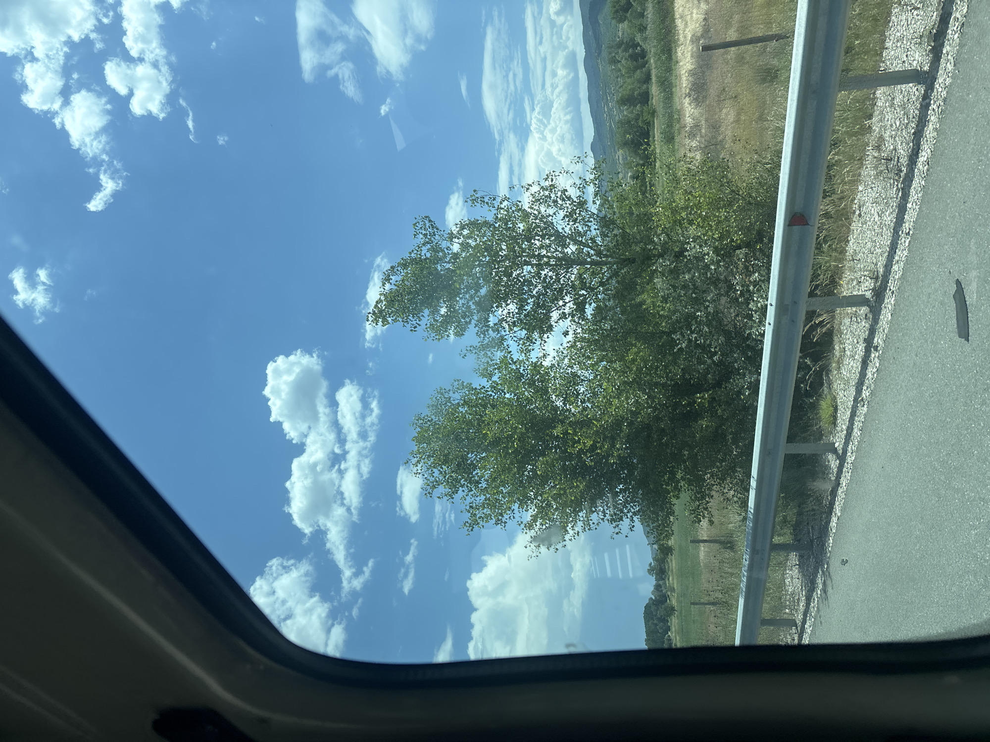 View from inside a vehicle through an open sunroof, looking towards a bright blue sky with scattered white clouds. To the right, roadside vegetation and a metal guardrail are visible. Further in the background, a landscape including fields and distant mountains is seen. The location is Strumyani, Bulgaria.