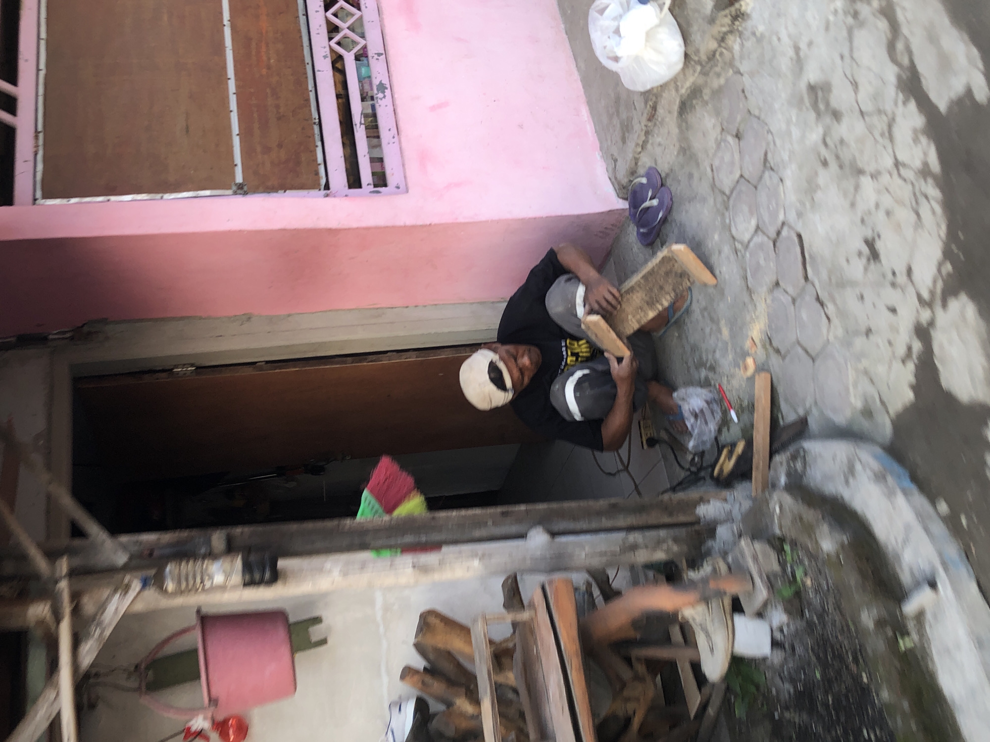 A man, wearing a black t-shirt, grey pants, and a beige baseball cap, is crouched on a concrete pathway in Keboguyang, Indonesia. He is working on a piece of wood, holding a rasp or file in his right hand and the wood in his left. A power tool is visible on the ground near his feet, along with a broom with red, green, and yellow bristles. To his right, a white plastic bag and a pair of purple flip-flops are on the ground. A pink building with a brown window is to his right, and a wooden structure with various objects, including a pink bucket, is to his left. The ground is a mix of concrete and hexagonal paving stones, with some debris and a water channel in the foreground.