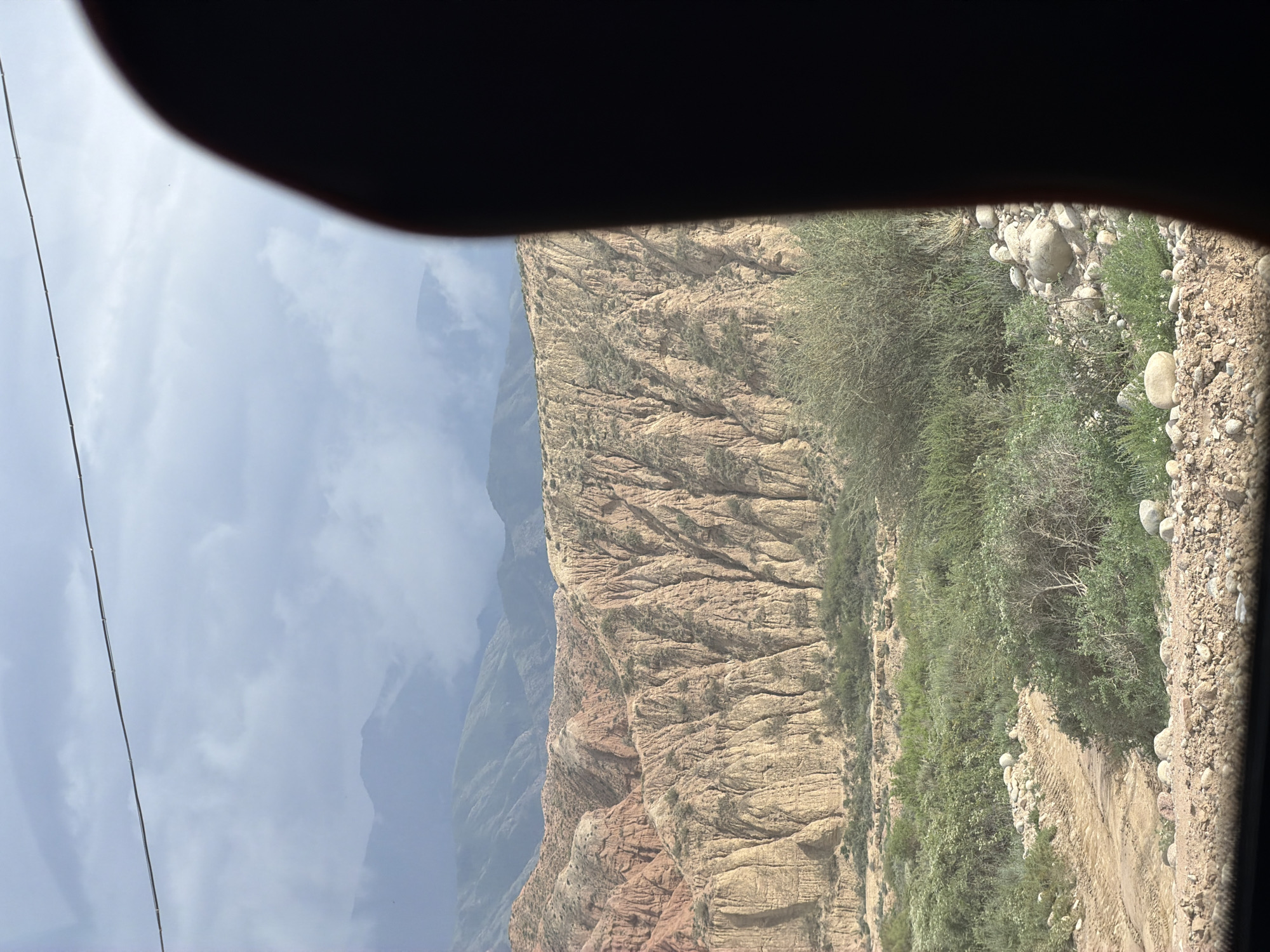 The image captures a scenic view of mountainous terrain, likely in or around Tong, Kyrgyzstan. In the foreground, a dirt road winds through a landscape dotted with green bushes and scattered rocks. The middle ground is dominated by a steep, eroded mountainside with distinct layers of reddish-brown and yellow rock formations, characterized by vertical striations and deep ravines. Sparse vegetation clings to the slopes. In the background, successive layers of mountains recede into the distance, partially obscured by a hazy, overcast sky. A dark, blurred object, possibly a person's hand or head, occupies the right side of the frame, partially obscuring the view of the sky and distant mountains. A thin, dark horizontal line, likely a wire, stretches across the upper portion of the sky.