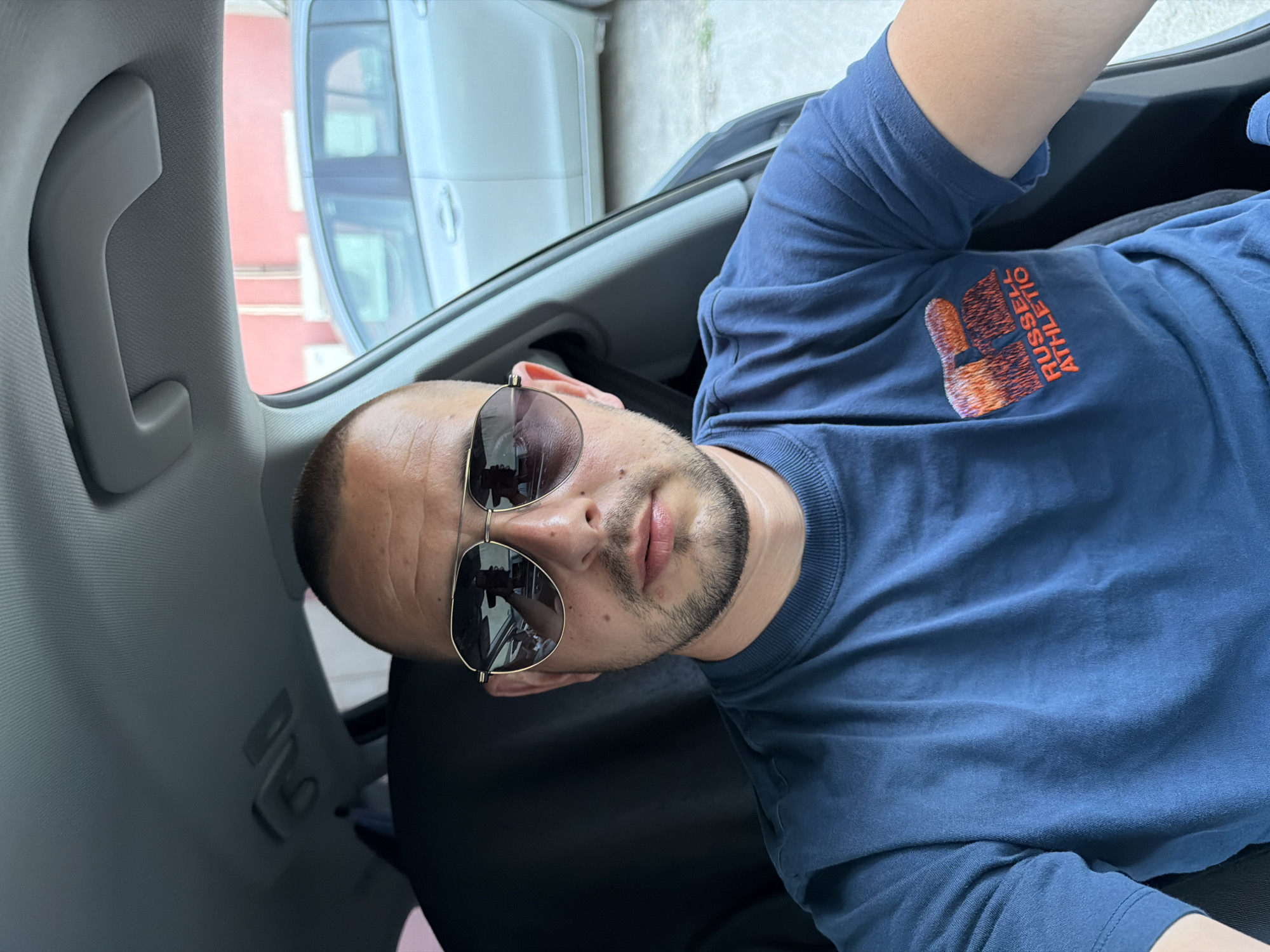 A man is sitting in the driver's seat of a car, looking at the camera. He is wearing a blue t-shirt with the Russell Athletic logo on it and aviator sunglasses. The interior of the car is visible, including the headliner and a passenger seat. Outside the car, another car and a building are visible. The lighting suggests it is daytime. The location is the city of Burgas, Bulgaria.