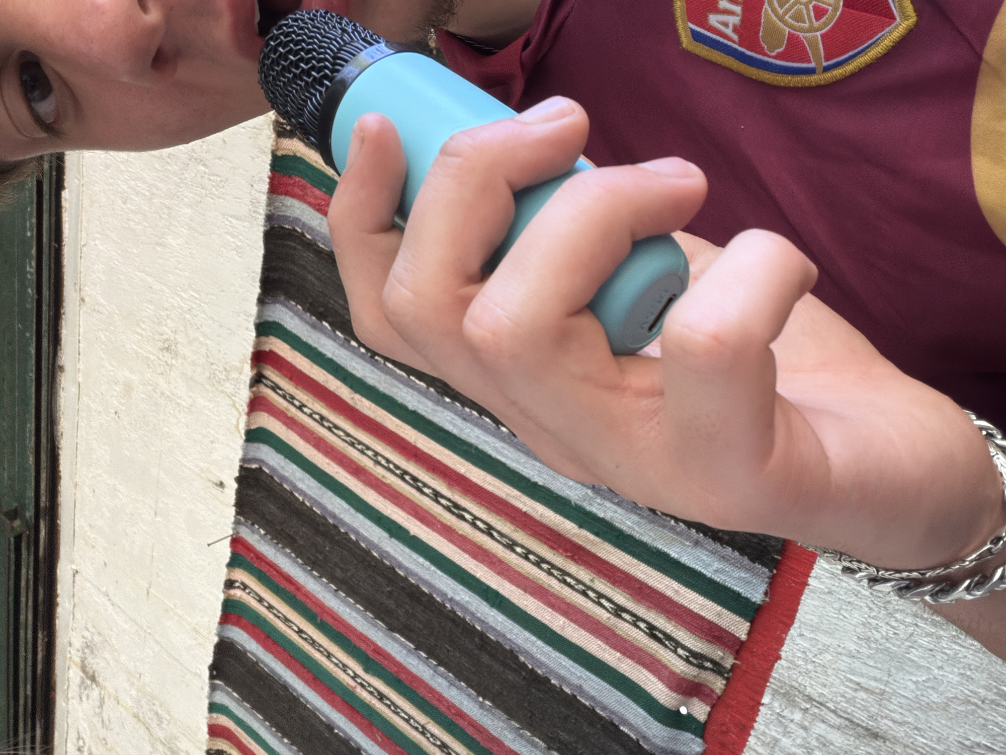 A person holds a blue microphone close to their mouth. The person is wearing a maroon shirt with a yellow and red patch on the upper right chest area and a silver bracelet on their right wrist. A striped rug with shades of gray, green, red, white, and black is partially visible. A white textured wall and a green door are partially visible on the left. The location is not identifiable as Kanchevo, Bulgaria from the visual information provided.