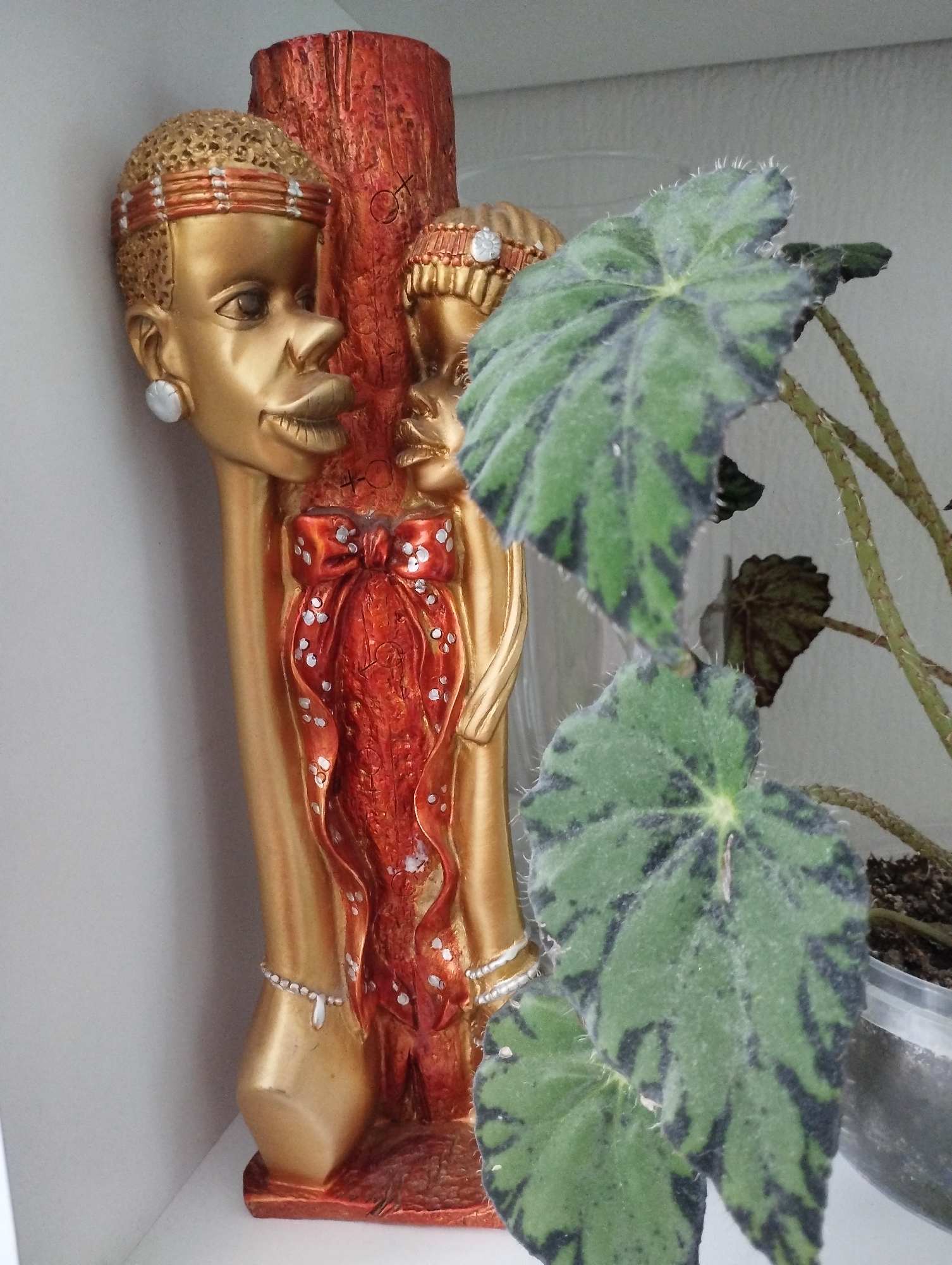 A medium, indoor shot features a tall, golden-colored statue of two African individuals, juxtaposed with a potted plant. The statue is adorned with what appears to be a red ribbon with white dots and white bracelets. The statue's red portion exhibits the symbol with the cross inside the circle. A green, textured plant, potentially a Begonia, with dark, mottled patterning on its leaves, is placed on the right. The plant is potted in a clear plastic container with visible soil. The setting appears to be a white shelf or surface in a room. Location details, like Sadovyy, Russia, are not ascertainable from the image alone.
