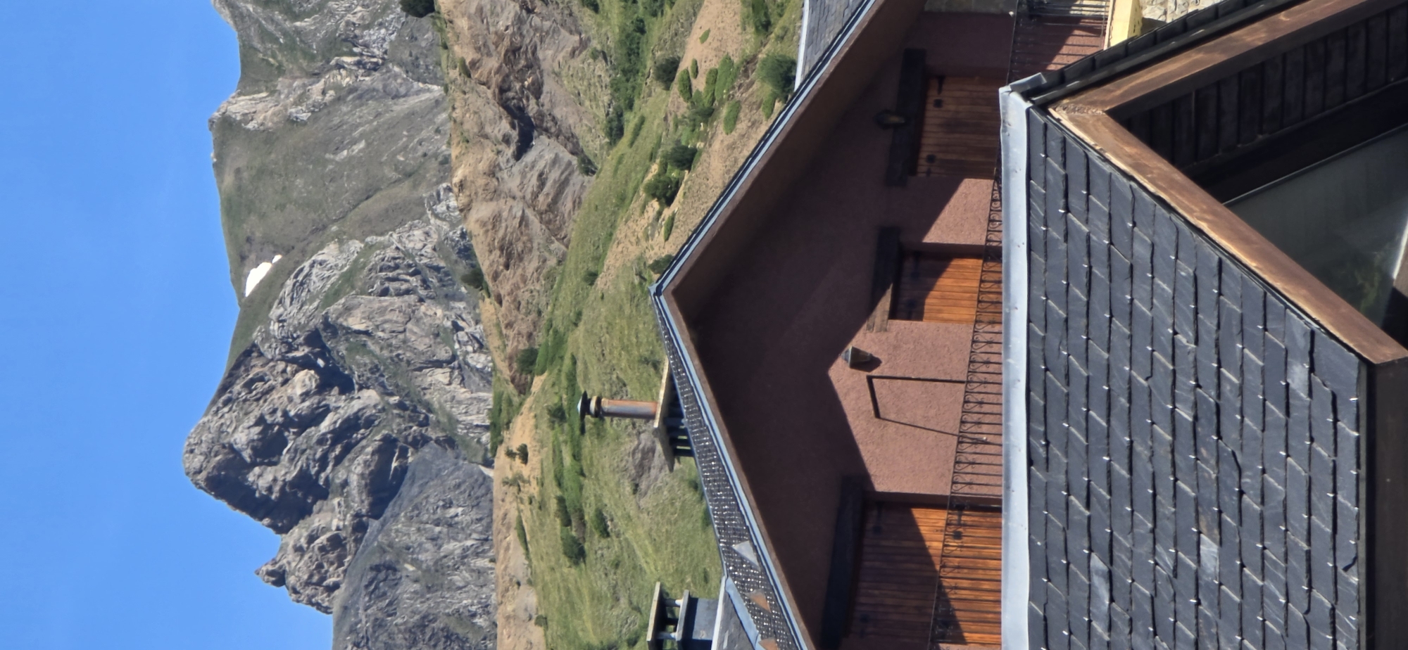 The image shows a close-up view of a building with a dark slate roof, situated against a backdrop of rugged, green mountains and a clear blue sky. The building's exterior is a warm, reddish-brown color, with dark wooden shutters or doors visible. A metal chimney pipe extends from the building. The mountains are rocky and steep, with patches of green vegetation and some remaining snow on the higher slopes. The sunlight suggests it is daytime, and the clear sky indicates fair weather. The scene evokes a sense of remoteness and natural beauty, typical of a mountain landscape. The image appears to be taken from a slightly elevated perspective, looking down at the building and the surrounding terrain. There is no visible text in the image.