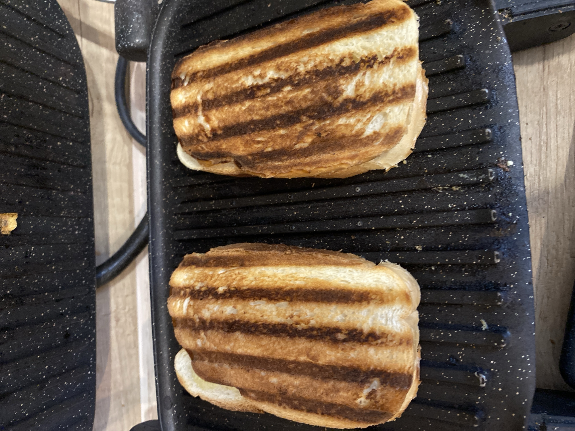 Two grilled cheese sandwiches are cooking on a black, ribbed electric grill. The sandwiches have golden-brown crusts with distinct grill marks and appear to be filled with melted cheese. The grill is placed on a light-colored wooden surface. The background shows the upper part of the grill with its textured black surface and a black cord extending from it. The scene does not provide any information to determine the location in the city of Tyumen, Russia.