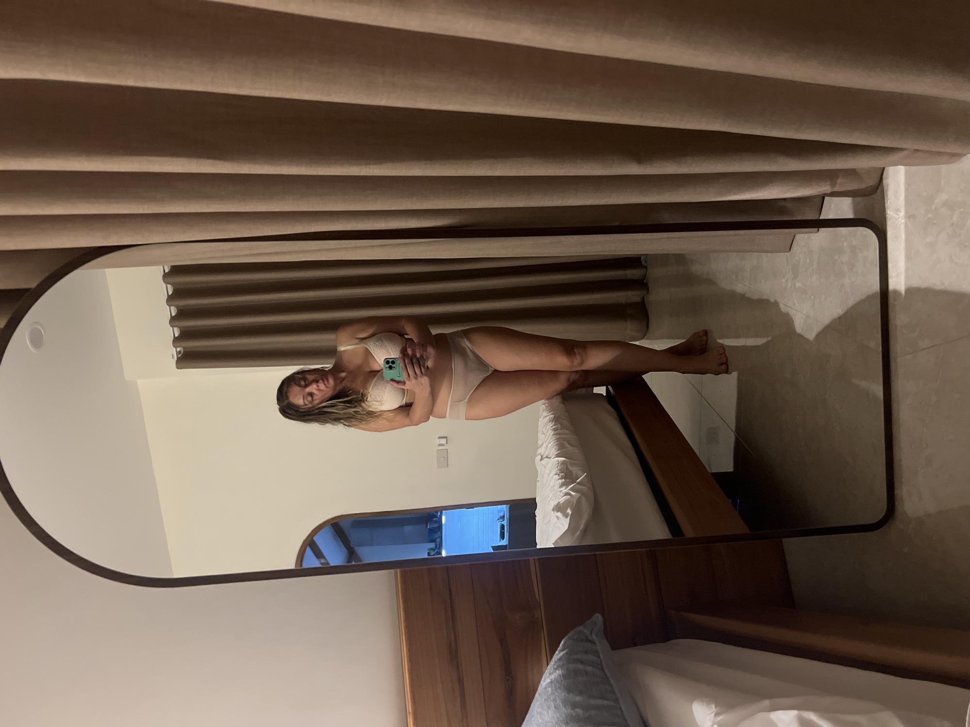 A woman is standing in front of a full-length mirror, holding a phone to take a selfie. She is wearing a beige bra and matching underwear. The mirror reflects a bedroom with a bed on the left and a window with curtains on the right. The room appears to be in a tropical location, likely in or near Koh Phangan, Thailand, suggested by the style of the curtains and the overall ambiance. The floor is tiled. The woman's reflection shows her looking down at her phone with a neutral expression.