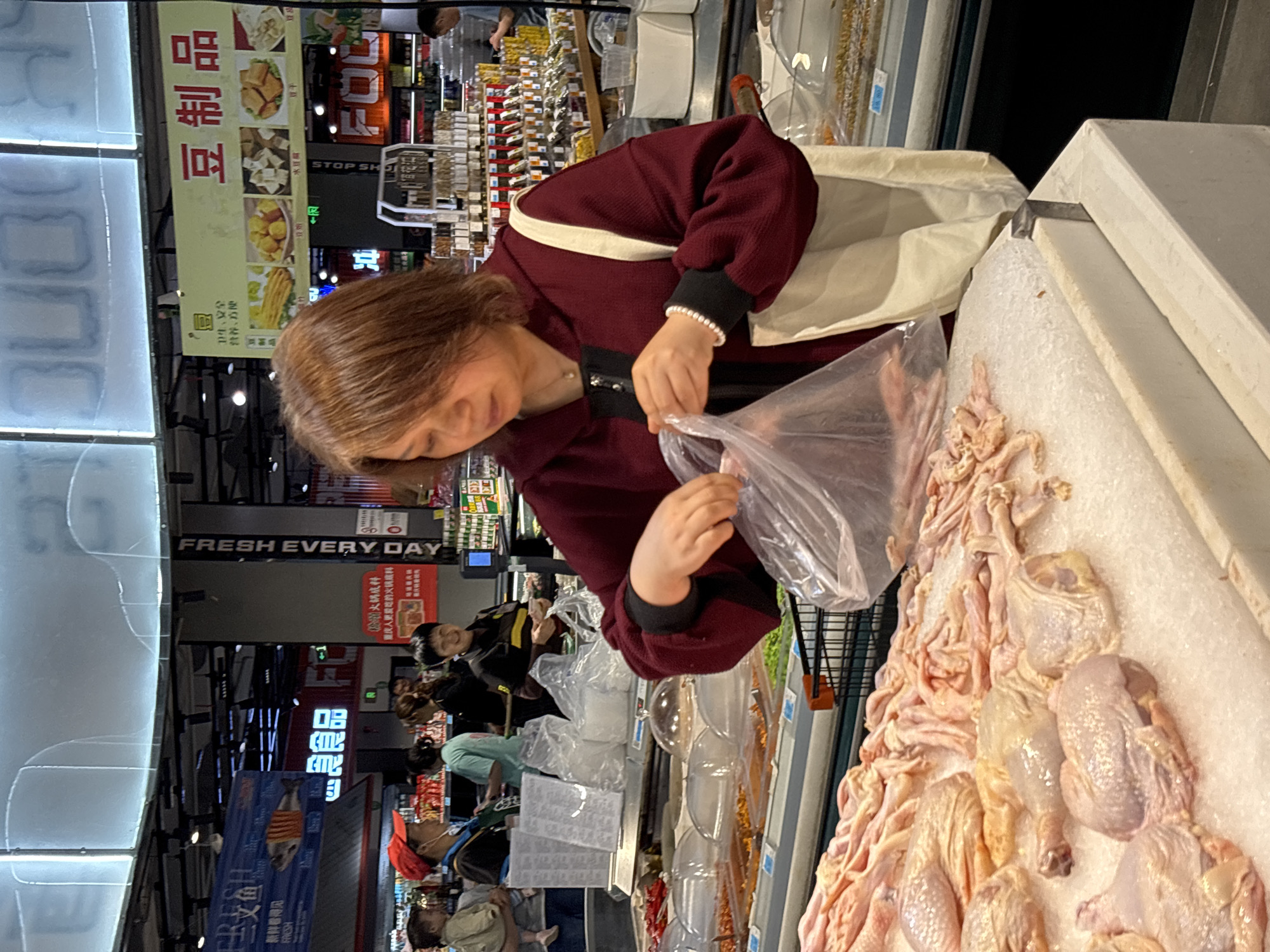A woman in a burgundy sweater and a white bracelet is putting raw chicken into a plastic bag in a grocery store. The chicken is displayed on a bed of ice. Other customers are visible in the background, along with various signs in Chinese. The environment includes signs for "Fresh Every Day" and "Dou Products", with one sign stating "We Love Chongqing Hot Pot." The text suggests the location could be a supermarket in Jiangjin, Chongqing, China.