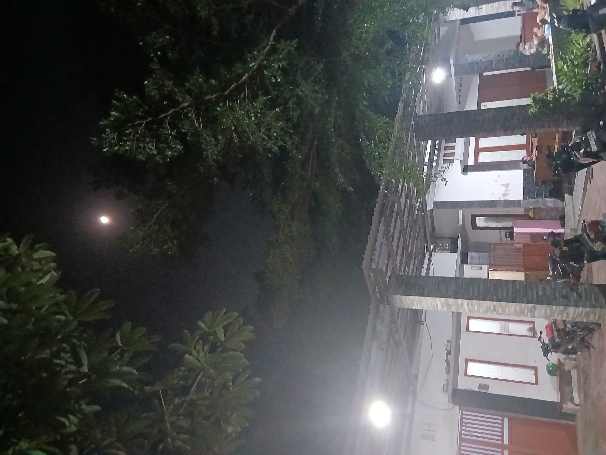 The image depicts a dimly lit outdoor scene at night, likely a residential area in Sekarputih, Indonesia. A full moon is visible in the dark sky, partially obscured by the branches of lush green trees that dominate the upper portion of the frame. Below the trees, a covered patio area of a building is visible, supported by stone pillars. The building appears to be white with prominent red doors and windows. Several motorcycles are parked in the foreground. In the background, a group of people are seated on a wooden bench, seemingly socializing. The overall atmosphere is relaxed and communal.