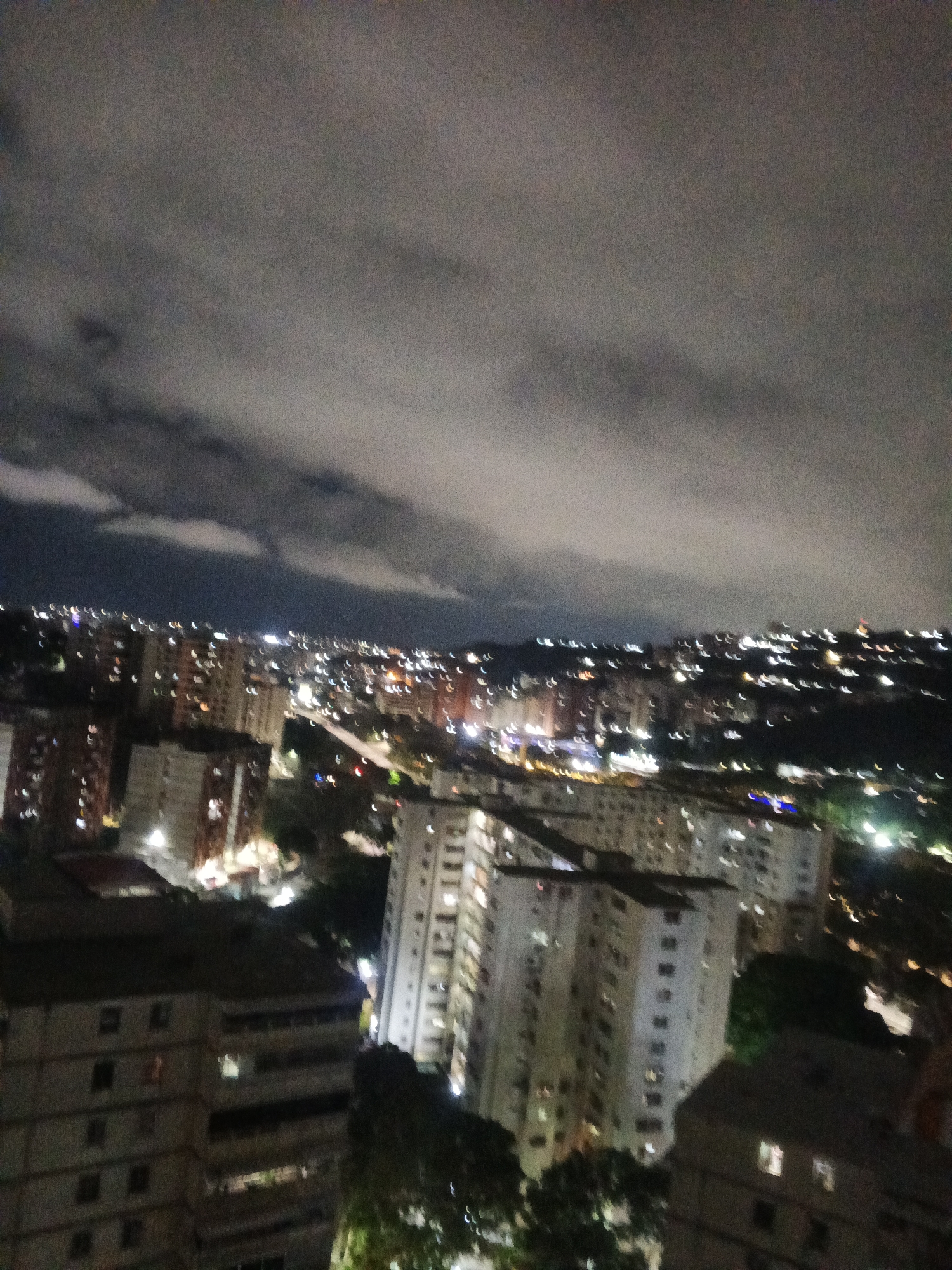 The image shows a nighttime view of Caracas, Venezuela, featuring numerous buildings illuminated by lights. The sky is overcast with clouds. The foreground shows residential buildings, and in the background, a cityscape with more lights indicating the populated area. The photo is somewhat blurry, suggesting it might have been taken in low light or with movement.