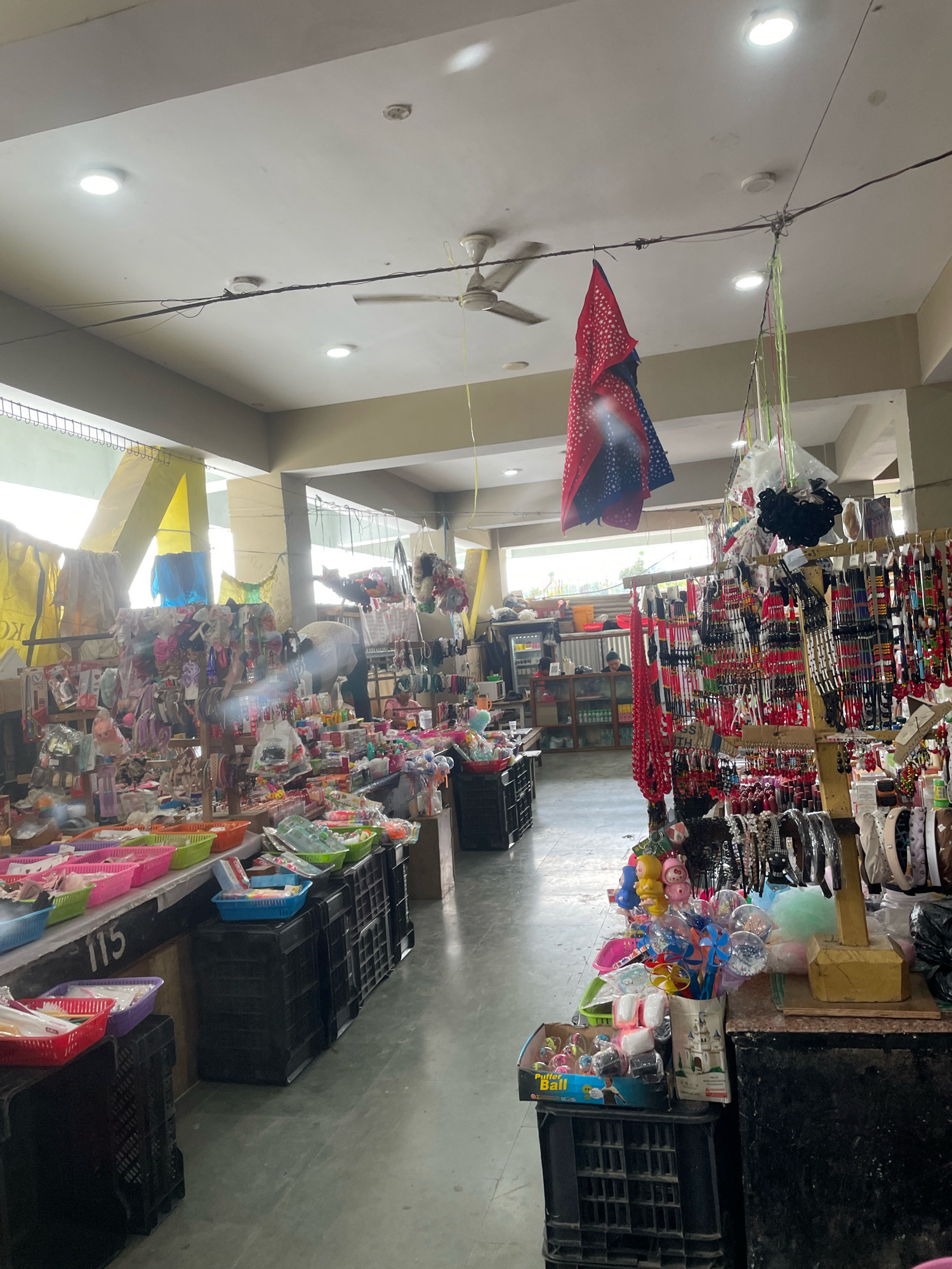 The image depicts a bustling market stall in Kangpokpi, India, filled with a variety of accessories and toys. The stall features numerous racks and shelves laden with colorful items, including beaded necklaces, headbands, hair clips, and small toys. In the foreground, several plastic crates are stacked, holding baskets filled with more merchandise. A red and blue patterned cloth hangs from a wire above, adding a splash of color to the scene. The market appears to be indoors, with a white ceiling and fluorescent lighting. A ceiling fan is visible in the upper portion of the image. The floor is a light-colored concrete or tile. Several shoppers are visible in the background, browsing the stalls. The overall impression is one of a vibrant and crowded marketplace.