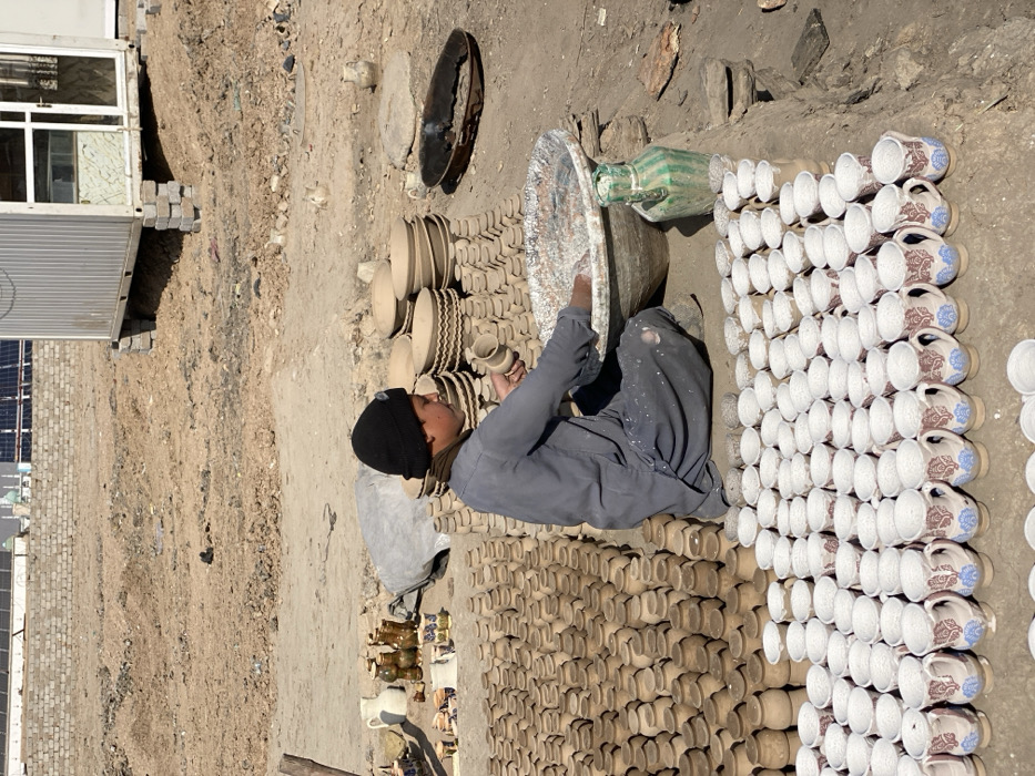 The scene depicts an outdoor pottery workshop in Kandahar, Afghanistan, under bright daylight. A young male, dressed in a grey tunic and dark pants, a black beanie, and with white splatters on his clothing, kneels on the dry, dusty earth. He is actively engaged in coating a small, unglazed clay cup by dipping it into a large, grey metal basin containing a white, viscous substance, likely slip or glaze.
Around him, numerous pottery items are arranged:
* In the immediate foreground, multiple rows of finished or semi-finished mugs with handles, decorated with blue and reddish-brown stylized floral patterns, are neatly aligned. Behind these are rows of smaller, plain white-coated cups.
* To the male's left, hundreds of unglazed, light-brown clay pots or jars are stacked in organized rows. Further left, a small collection of decorated vases, a white pitcher, and other pottery pieces are displayed on a mat.
* To the male's right, several stacks of larger, unglazed clay bowls, some featuring textured or patterned rims, are present. A tall, green-striped ceramic jug sits beside the large basin.
* A large, dark, shallow metal pan containing dark residue, possibly a firing tray or part of a makeshift kiln, rests on the ground to the right.
The background consists of an arid, uneven dirt landscape. A corrugated metal shipping container, modified with windows and resting on concrete blocks, is visible in the mid-ground. Behind it, a concrete block wall is present. Further in the distance, structures with solar panels on a rooftop and a faint green-domed building, characteristic of a mosque, are visible, indicating an urban setting. A grey sack lies near the displayed pottery items.