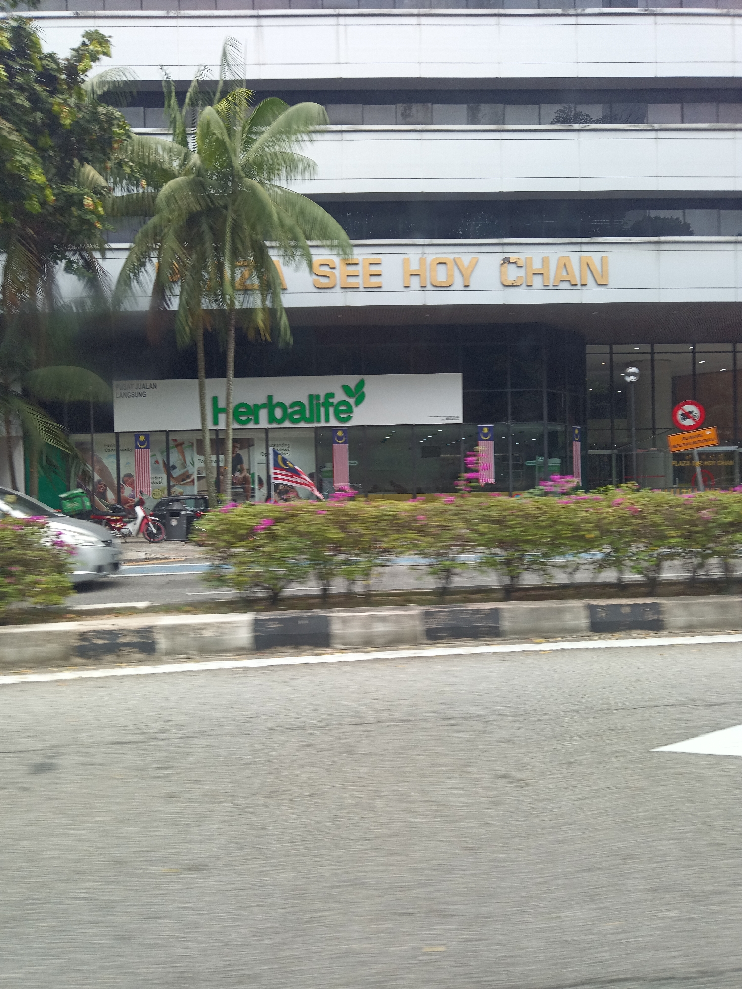 The image shows a street view in Kuala Lumpur, Malaysia, featuring the Herbalife building. A white building with horizontal window bands is visible in the background. The building has prominent gold lettering that reads "SEE HOY CHAN". Below this, a white facade displays the Herbalife logo and name in green. Malaysian flags are displayed in front of the building. In the foreground, a blurred street scene shows a silver car, a motorcycle, and lush green bushes with pink flowers. The road surface and a curb are also visible. The image appears to be taken from a moving vehicle, indicated by motion blur.