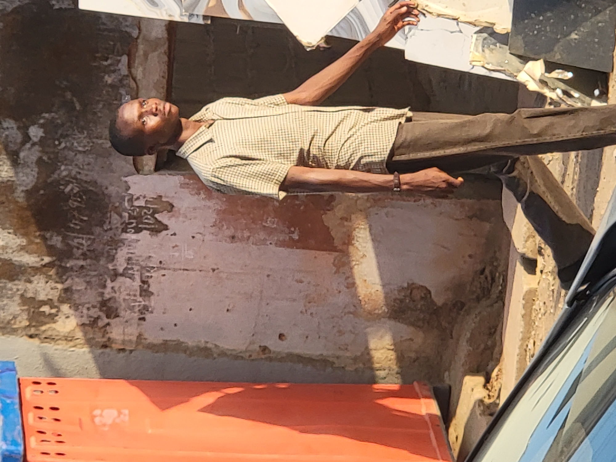 A man stands near a building in Maiburiji, Nigeria. The building has a textured, weathered facade with patches of dark and light coloration. The man is wearing a short-sleeved, checkered shirt and dark pants. He has a watch on his left wrist. He has his left arm raised. There are building materials and possibly tiles above him. At the bottom of the image is a blue crate and an orange plastic container. Part of a car is visible to the right.