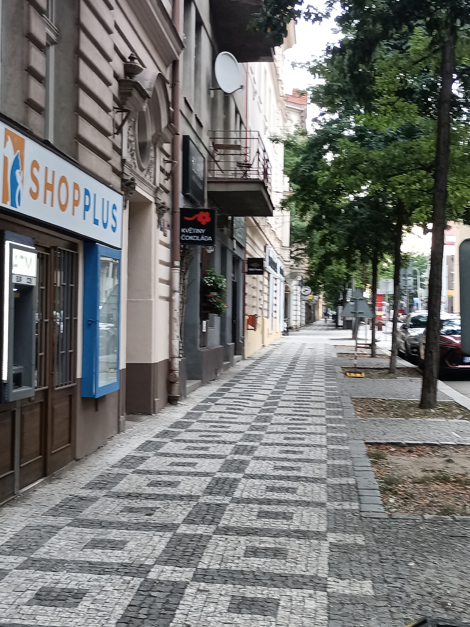 The image depicts a street scene in Prague, Czechia, likely in a commercial district. The perspective is from a sidewalk that is paved with a distinctive black and white patterned cobblestone design. On the left side of the image, a storefront with the sign "SHOPPLUS" is visible, featuring a blue window frame and an ATM machine. Adjacent to this is another shop with a dark sign that appears to be for a flower and chocolate shop, indicated by the text "KVĚTINY ČOKOLÁDA". The buildings lining the street have a traditional European architectural style with ornate details. Trees with full green foliage are planted along the sidewalk, providing shade. The street is lined with parked cars, and further down the road, other buildings and trees are visible. The overall atmosphere suggests a daytime scene in an urban environment.