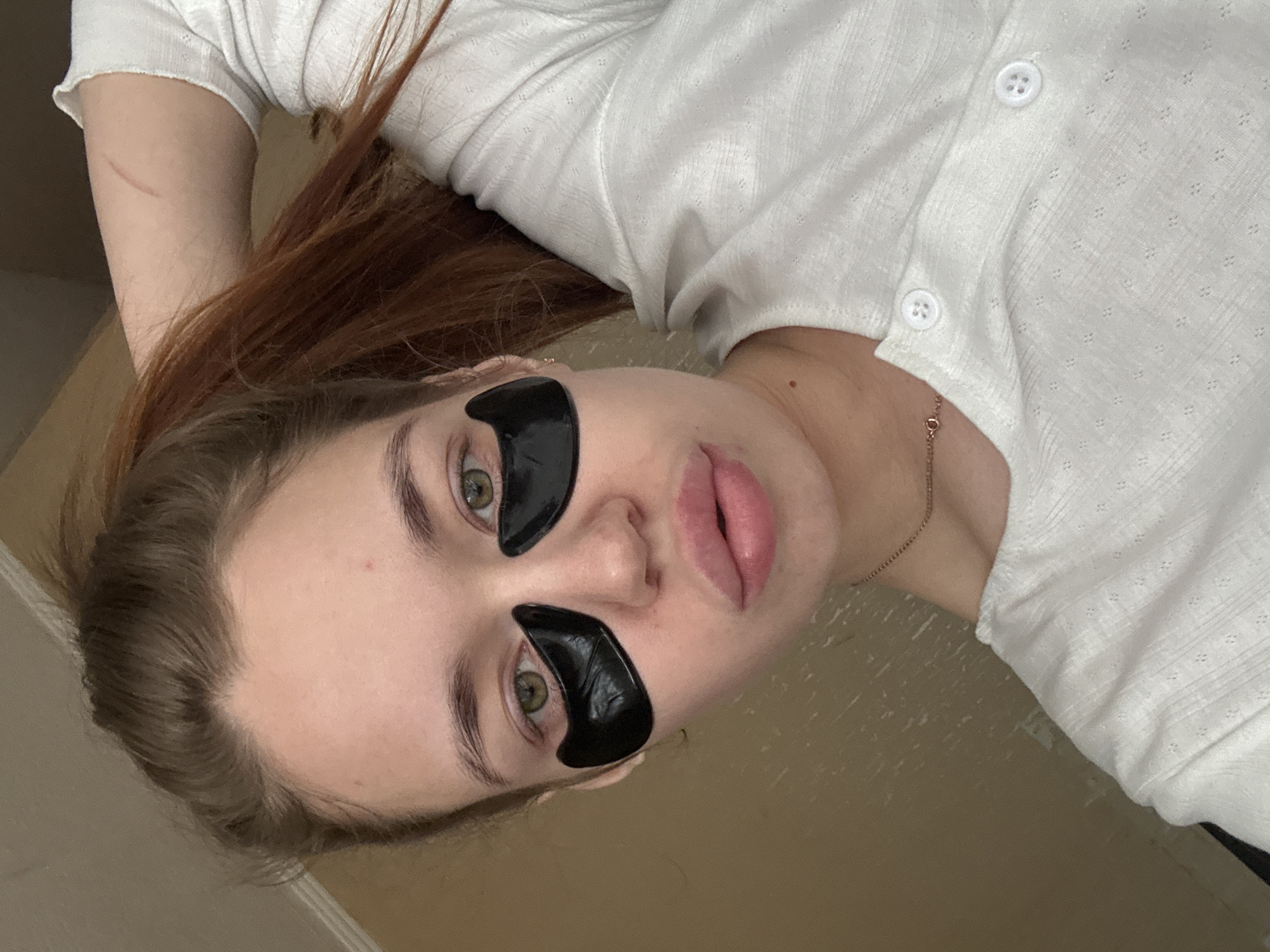 A woman lies on her back. She is wearing two black under-eye patches and a white shirt. She has brown hair, fair skin, and light green eyes. A gold necklace is visible around her neck. The background appears to be a neutral-colored wall. The location is unknown, but is potentially Chaikovsky, Russia.