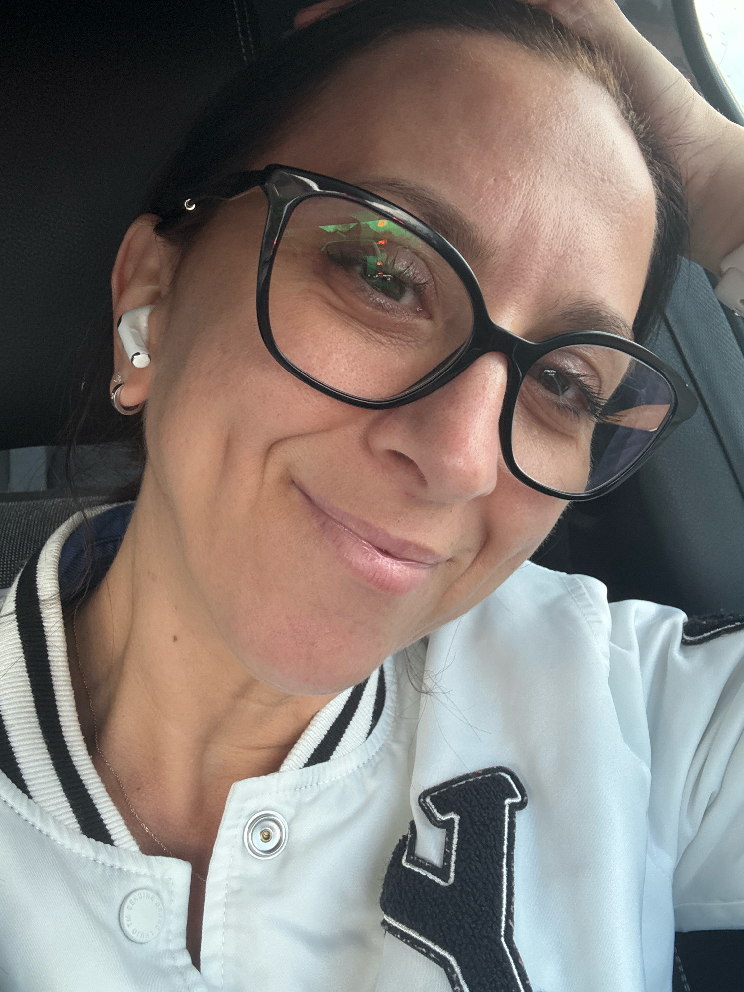 A woman with dark hair and glasses is taking a selfie. She is wearing a white jacket with black and white striped collar. She has an earbud in her right ear and a small hoop earring in her left ear. The background appears to be the interior of a vehicle. The image does not provide any specific information about the location within the city of Marum, Netherlands.