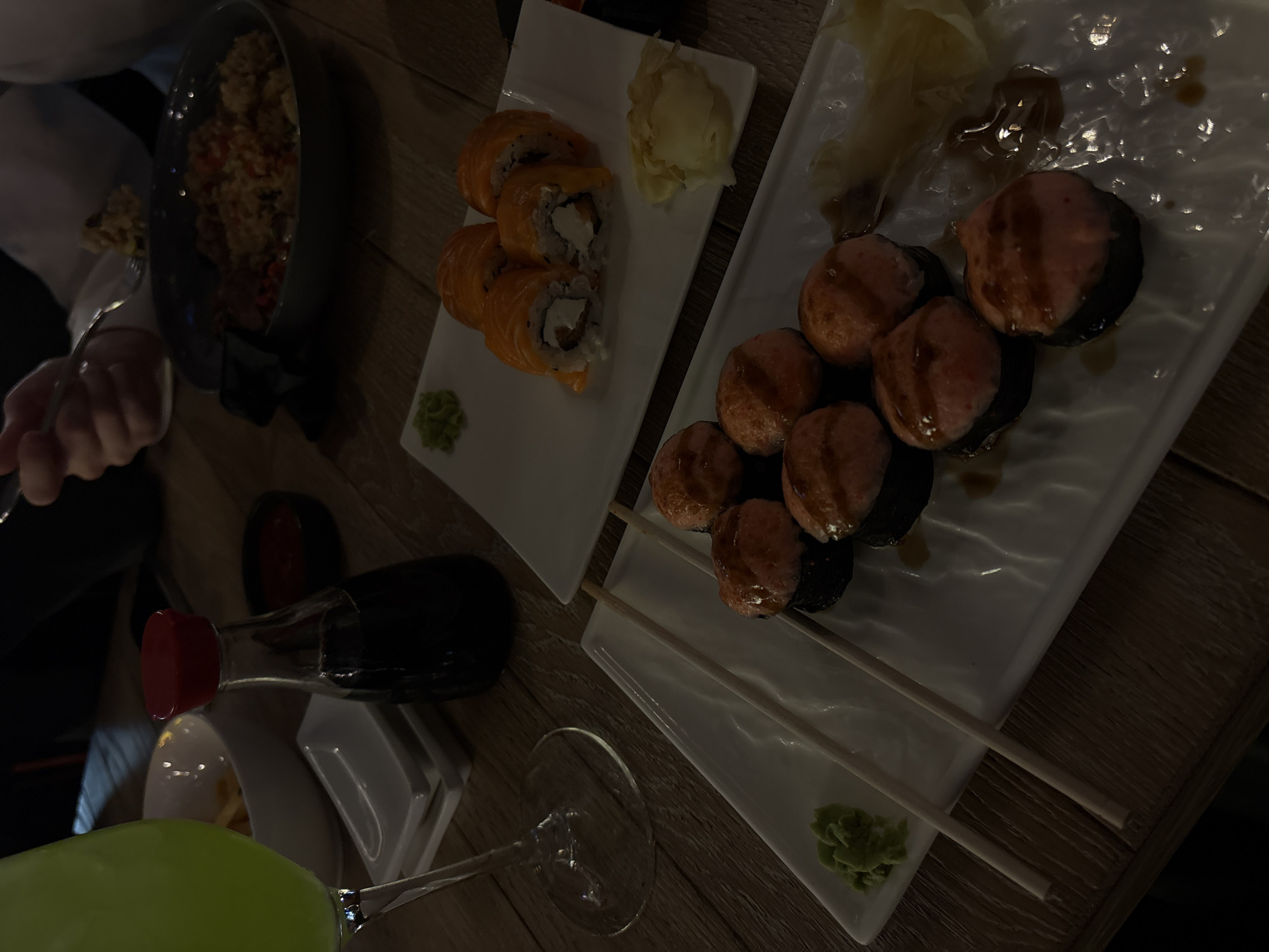 The image shows a table with several dishes of food, including sushi and what appears to be rice in a bowl. There is a person visible in the upper left, holding a fork. Also on the table are soy sauce, a glass, chopsticks, and condiments. The location is Unknown, Russia.