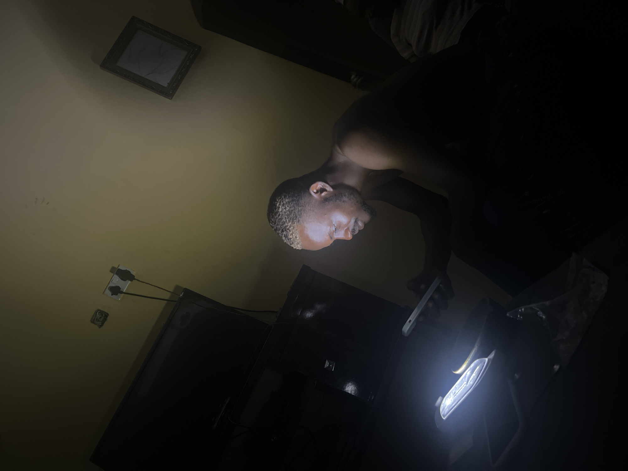 A man with short blonde hair, stands shirtless in a darkened room illuminated by the light of a mobile phone. The man appears to be inspecting a black suitcase with a silver lock. Behind the suitcase, a clear plastic bag with contents obscures the background. A TV is visible to the left. A picture hangs on the wall, above a dual electric outlet. Given the prompt, this scene purportedly takes place in Hanwa, Nigeria.
