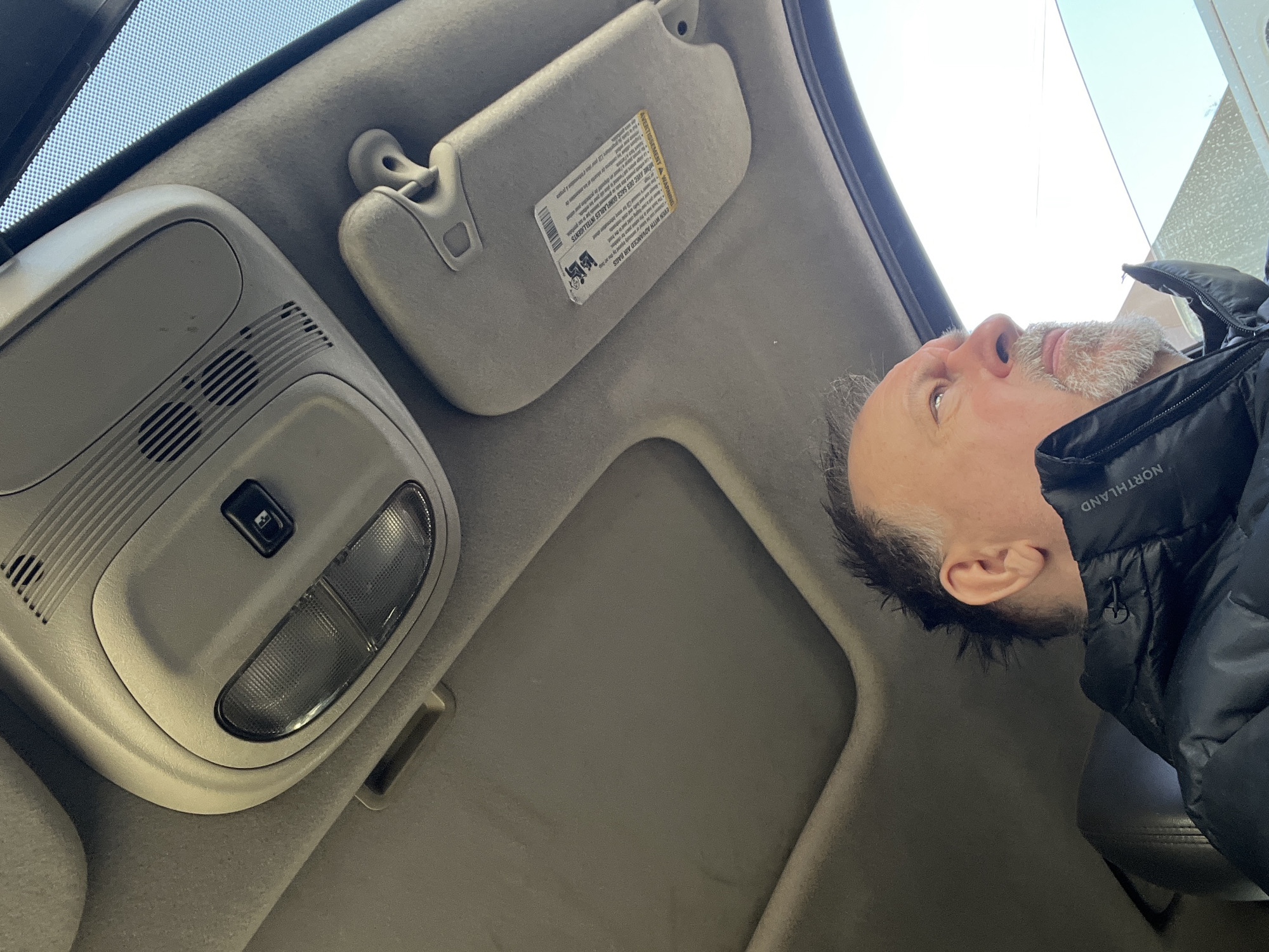The image shows the interior of a vehicle, likely a car, with a man looking upwards. The interior is fitted with an overhead console featuring a light and other controls, as well as a sun visor. The man appears to be in his late middle age, with graying hair and a beard. He is wearing a dark-colored, puffy jacket. The environment visible through the windshield suggests an outdoor setting with sky and possibly buildings. There is no information to support the claim that this is Podolsk, Russia.